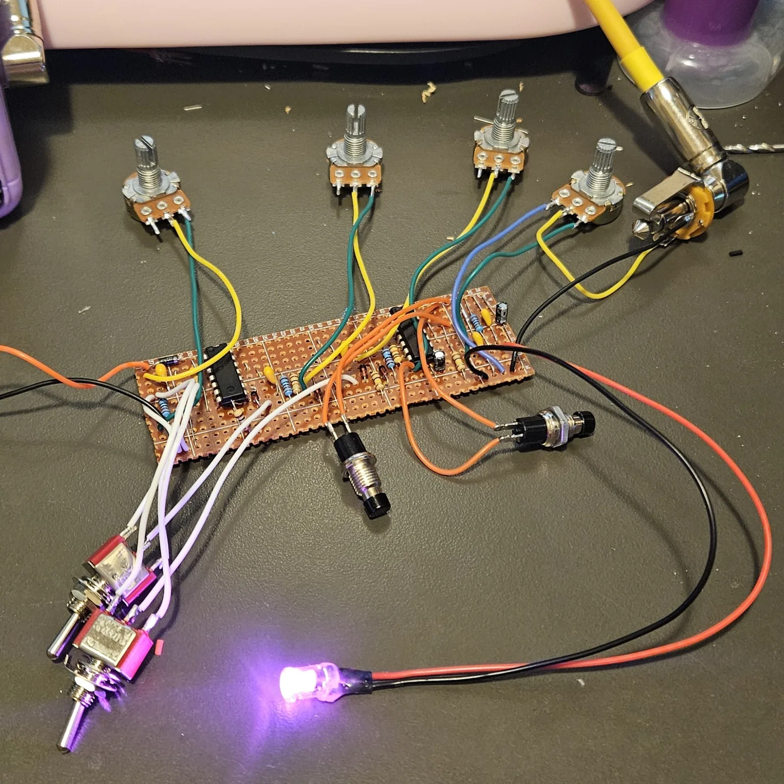 The finished board on a dark desk mat, wired up but not yet boxed. A rectangle of stripboard lies in the middle, copper side up, with two black chips in sockets of solder, small resistors and yellow and gray capacitors between them. Four potentiometers with bare knurled shafts and no knobs sit above it on yellow, green and blue wires, two black pushbuttons stand on orange wires in front of it, and two toggle switches with purple and pink bodies lie at the lower left on white and lilac wires. A pink LED is lit at the bottom on red and black leads, casting pink light across the mat. Orange and black supply wires run off to the left and a knurled hand drill lies at the upper right.