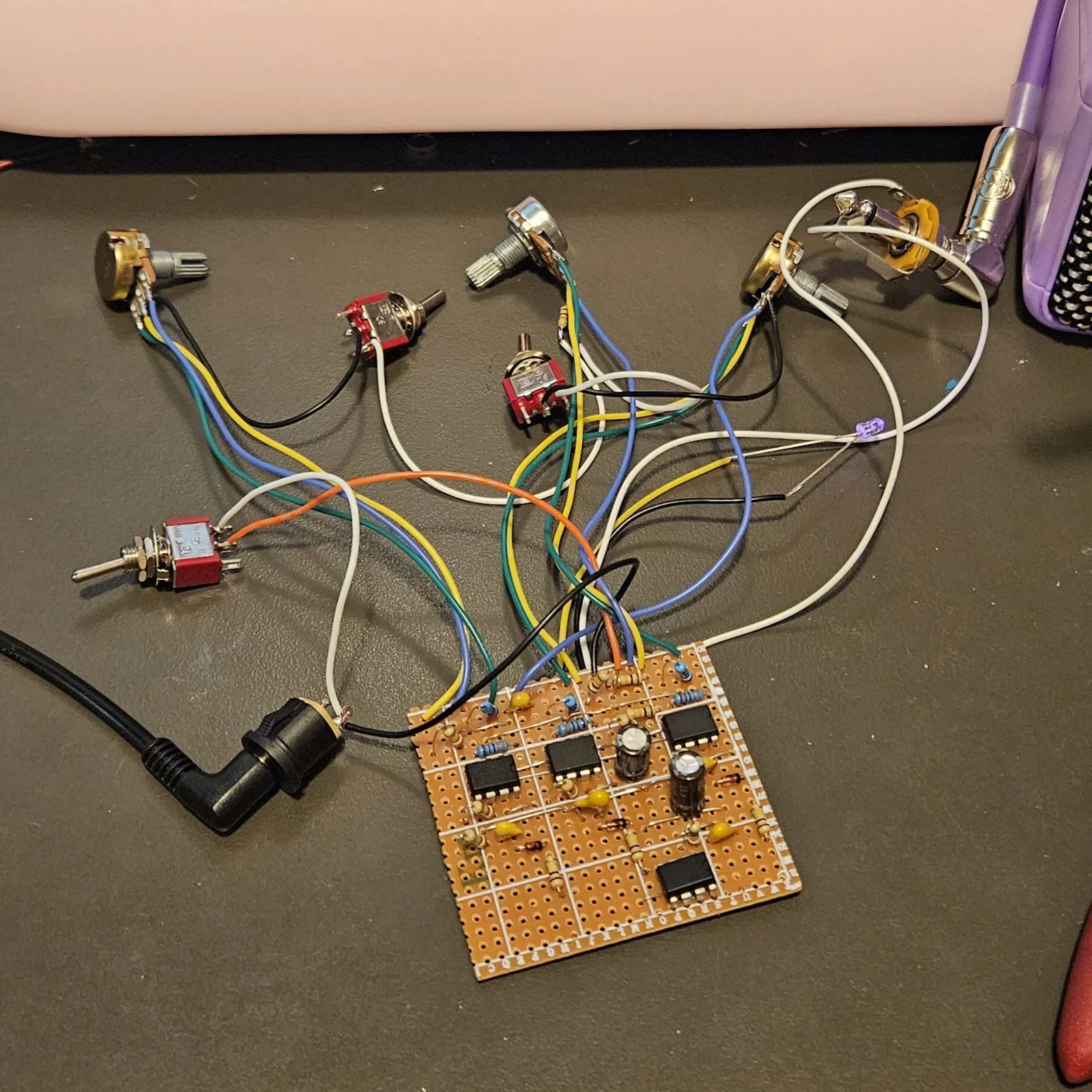 The finished board and all its off-board wiring spread out on a dark desk mat. At the bottom right, a square of tan stripboard with its row letters printed down the edge carries four small black eight-pin chips, blue and beige banded resistors, yellow disc capacitors and two black electrolytic cans. A bundle of orange, yellow, green, blue, black and white wires leaves its top edge and fans out across the mat to six potentiometers and three red toggle switches. Two small beige resistors are soldered inline in the wiring near the middle pot, joining it toward two of the switches. A violet LED burns in one of the white wire runs on the right. A black right-angle DC plug enters at the lower left, and a quarter-inch jack at the top right runs off to the edge of a purple practice amp.