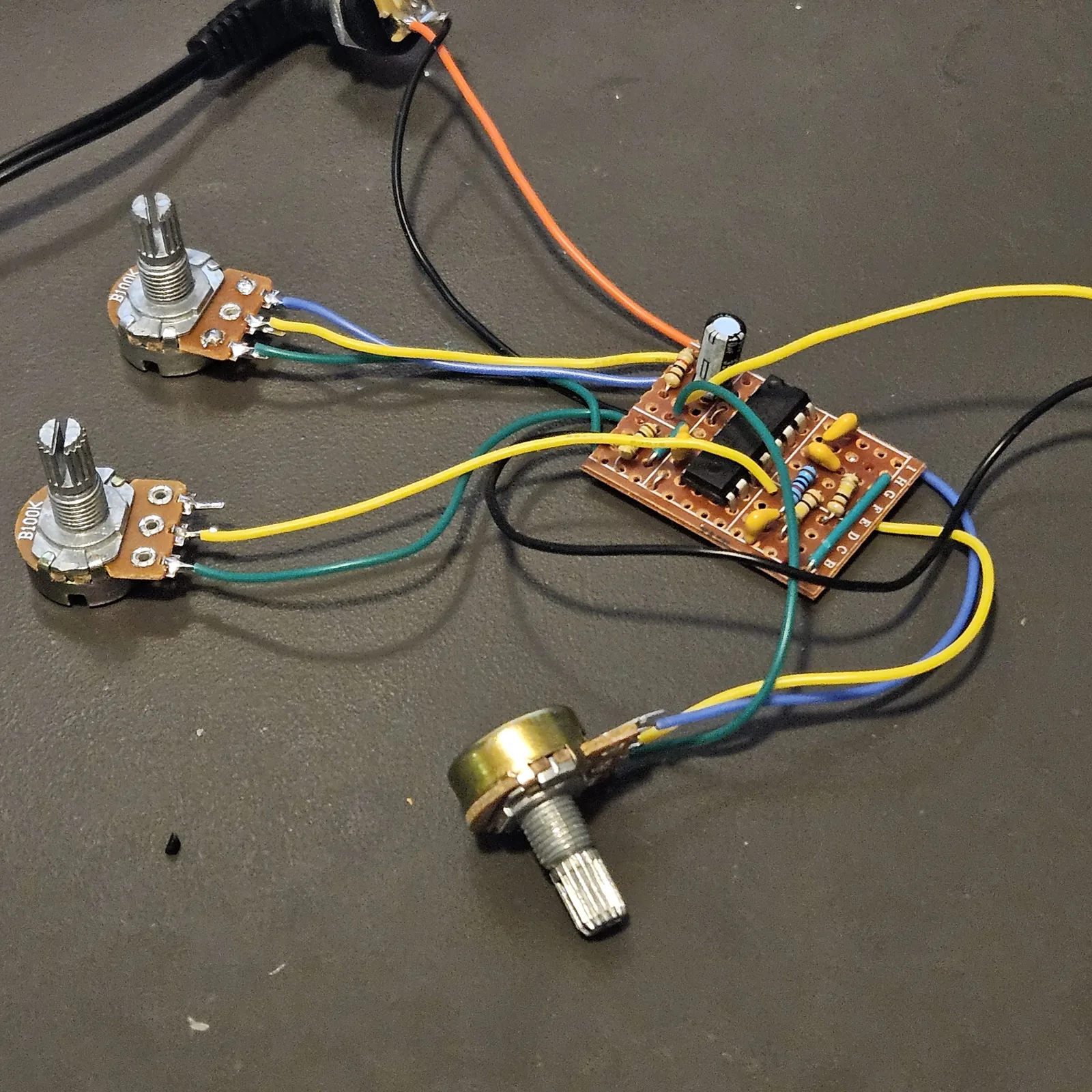 The 9 by 14 board wired up loose on a dark gray desk mat, not boxed. The small piece of stripboard sits at the right of center, its strips lettered B to I along one edge, with a black chip across the middle, a few beige and blue resistors, three yellow ceramic capacitors, a small electrolytic capacitor standing at the top edge and a green wire link. Blue, yellow and green wires run from the board to three pots with bare shafts: two marked B100K at the left, one above the other, and a third, brass-bodied, lying at the bottom. An orange and a black wire run from the top of the board up to a panel DC jack with a black power cable plugged in, and more black, yellow and blue wires leave the board to the right, out of the frame. There is no toggle switch in the picture.