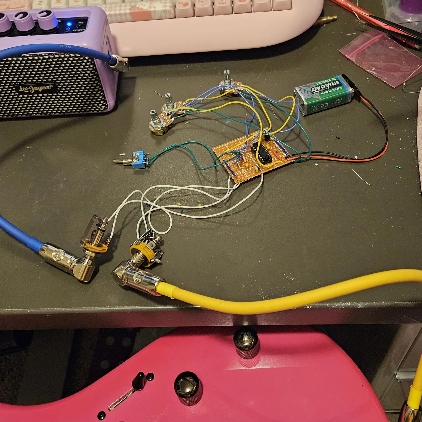 The finished board on a dark desk, wired but not boxed. A small rectangle of orange-brown stripboard sits in the middle with a 16-pin chip, a handful of resistors and small yellow capacitors, and blue wire links. Three potentiometers with bare shafts lie above it on blue and yellow wires, a small blue toggle switch lies to the left on green wires, and a green 9V battery sits to the right on red and black leads. Two quarter-inch jacks on white wires lie at the bottom left: one has a blue cable plugged in that runs up to a small lilac practice amp with its power light on, the other a yellow cable that runs down to the body of a pink electric guitar at the bottom of the frame. A pink keyboard sits behind the amp.