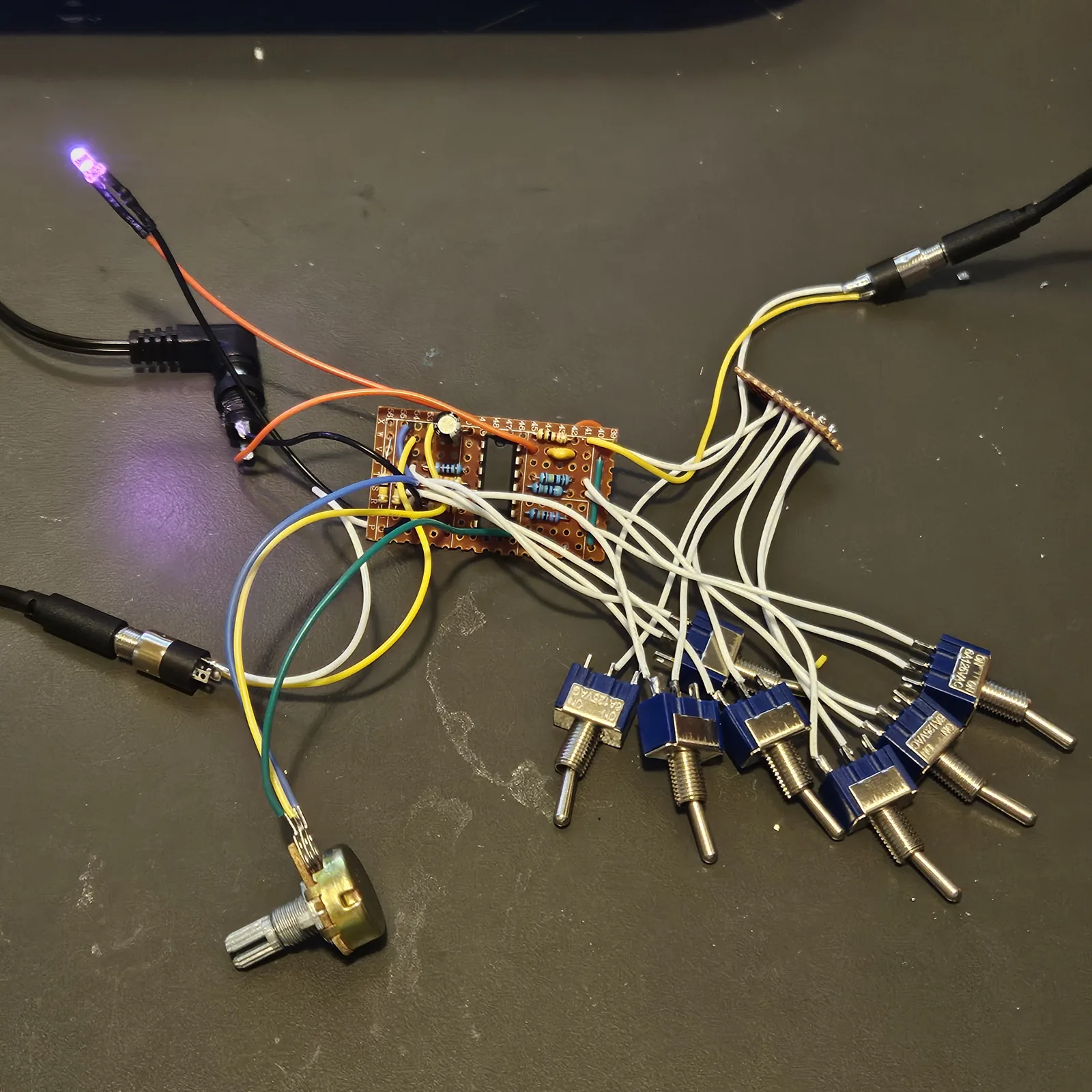 The 9 by 17 board wired up loose on a dark desk mat, not boxed. The small piece of stripboard sits in the middle with the CD4024 standing in its center, blue resistors lying along the strips either side of the chip, a small electrolytic capacitor at its top edge, a yellow ceramic capacitor and two wire links, one blue and one green. A bundle of white wires runs from the board down and to the right to seven blue miniature toggle switches with chrome levers, lying loose, and up to a narrow offcut of stripboard at the upper right where the white wires gather. Blue, yellow and green wires run down to a pot with no knob at the lower left. A right-angle power plug sits in a panel DC jack at the left on orange and black wires, black cables are plugged into a panel jack at the lower left and an inline jack at the upper right, and an LED on red and black leads is lit at the top left.