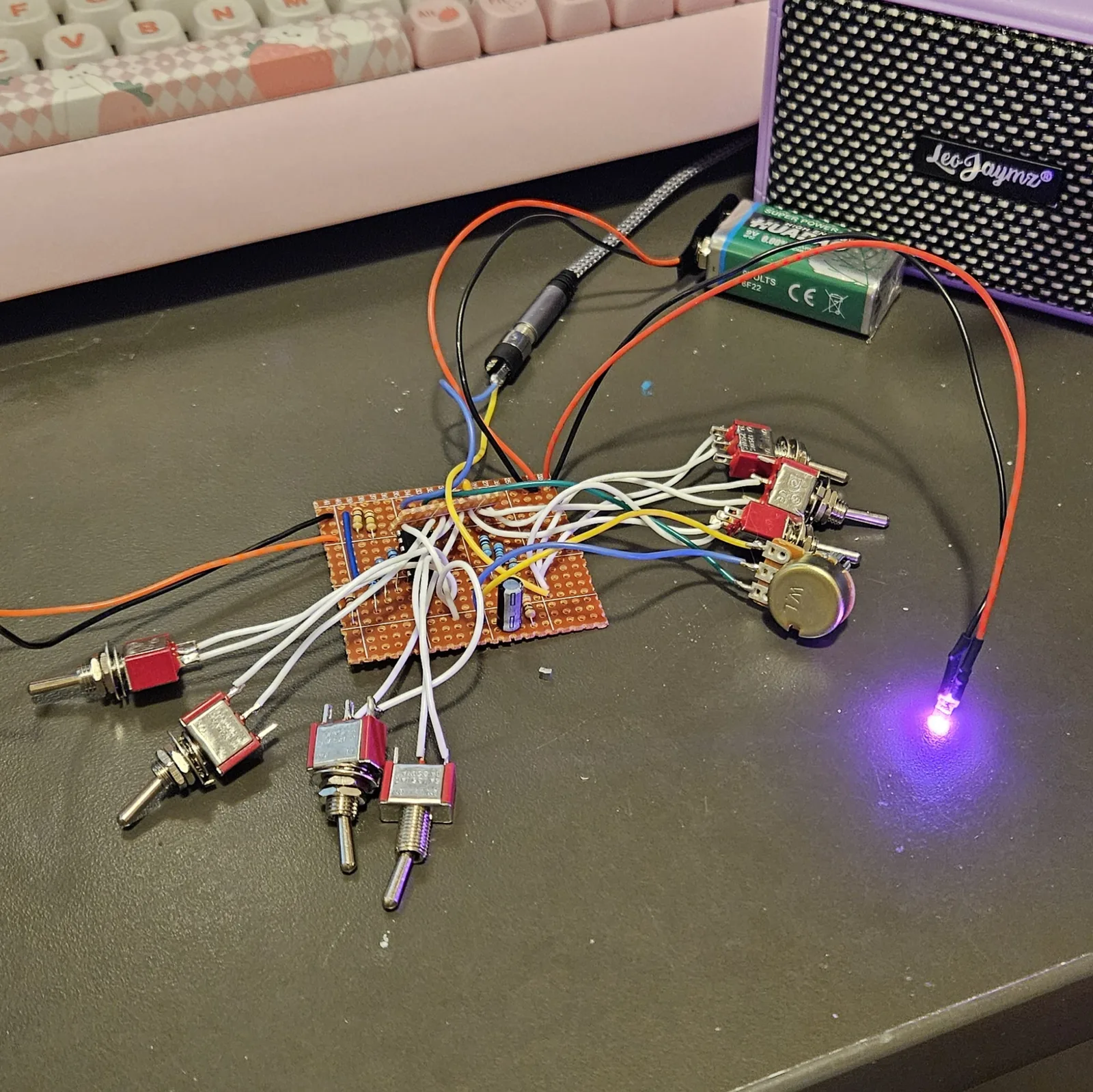The finished board on a dark desk, wired but not boxed. A small rectangle of orange stripboard sits in the middle with the CD4024, a row of resistors, one black electrolytic capacitor and two blue link wires. Seven small toggle switches with red bodies hang off the board on white leads, one to the left, three lying along the bottom of the frame and three standing to the upper right beside a volume pot on blue, yellow and green wires. A green 9V battery sits behind the board on red and black leads, a black cable runs from the board up to a purple practice amp lettered LeoJaymz at the top right, and a lit LED on red and black leads lies at the right. A pink keyboard sits behind.