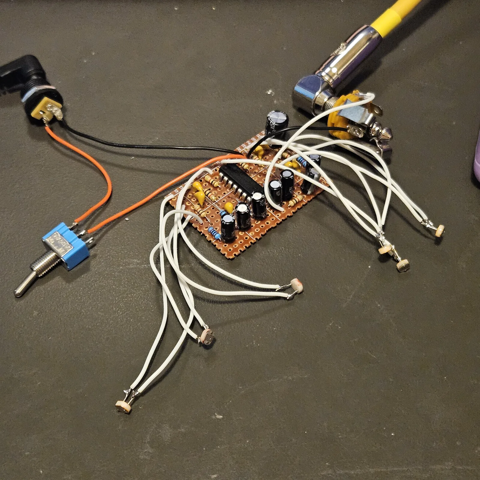 The 13 by 19 board wired up loose on a dark gray desk mat, not boxed. The small piece of orange stripboard sits in the middle with the CD40106 across it, black electrolytic capacitors standing around it, and small yellow ceramics and beige resistors. Twelve white wires fan out from it in pairs to six photocells lying loose on the mat, one at the end of each pair. At the left an orange and black pair runs to a small blue toggle switch and a DC jack, and at the top right a quarter-inch jack has a yellow cable plugged in.