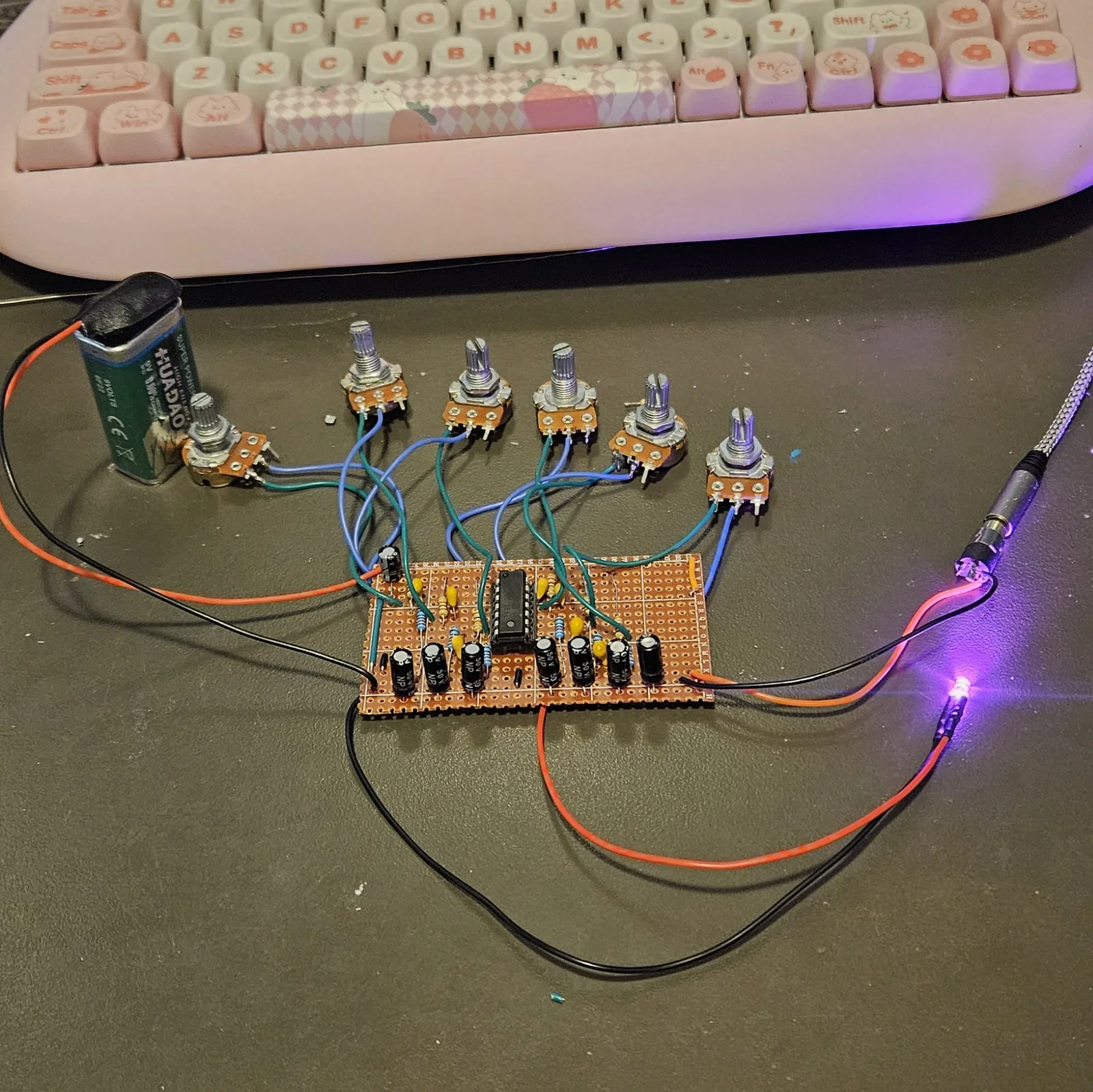 The finished board on a dark desk, wired but not boxed. A rectangle of orange stripboard sits in the middle with the CD40106 in the center, a row of seven black NP electrolytic capacitors along its bottom edge, another at the top left, and small yellow ceramics and resistors around the chip. Six pots stand in a row above it on blue and green flying leads, two wires each. A green 9V battery stands at the left on red and black leads, a braided cable with a jack plug lies at the right, and a lit LED on red and black leads sits at the lower right. A pink keyboard fills the top of the frame.