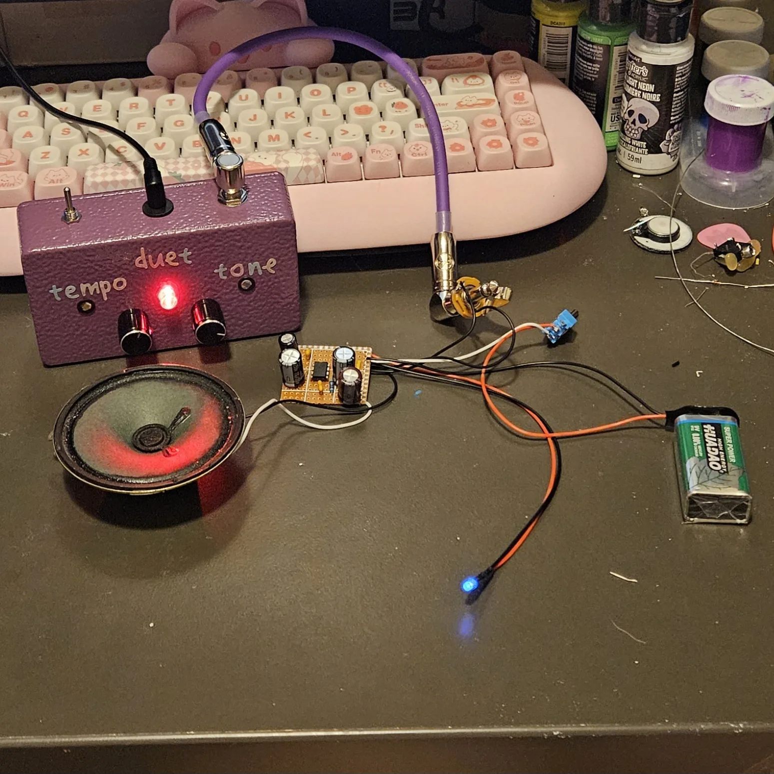 The finished board on a dark desk, wired but not boxed. A small rectangle of orange stripboard sits in the middle with the LM386, four black electrolytic capacitors and a few small parts. A black speaker lies to its left on white wires, its cone lit red by the LED of a purple pedal behind it lettered duet, tempo and tone, whose purple cable runs down to a quarter-inch jack on flying leads beside the board. A small blue toggle switch hangs off three leads to the right, a green 9V battery sits further right on red and black leads, and a lit LED on its own two leads lies at the bottom of the frame. A pink keyboard and jars of paint sit behind.