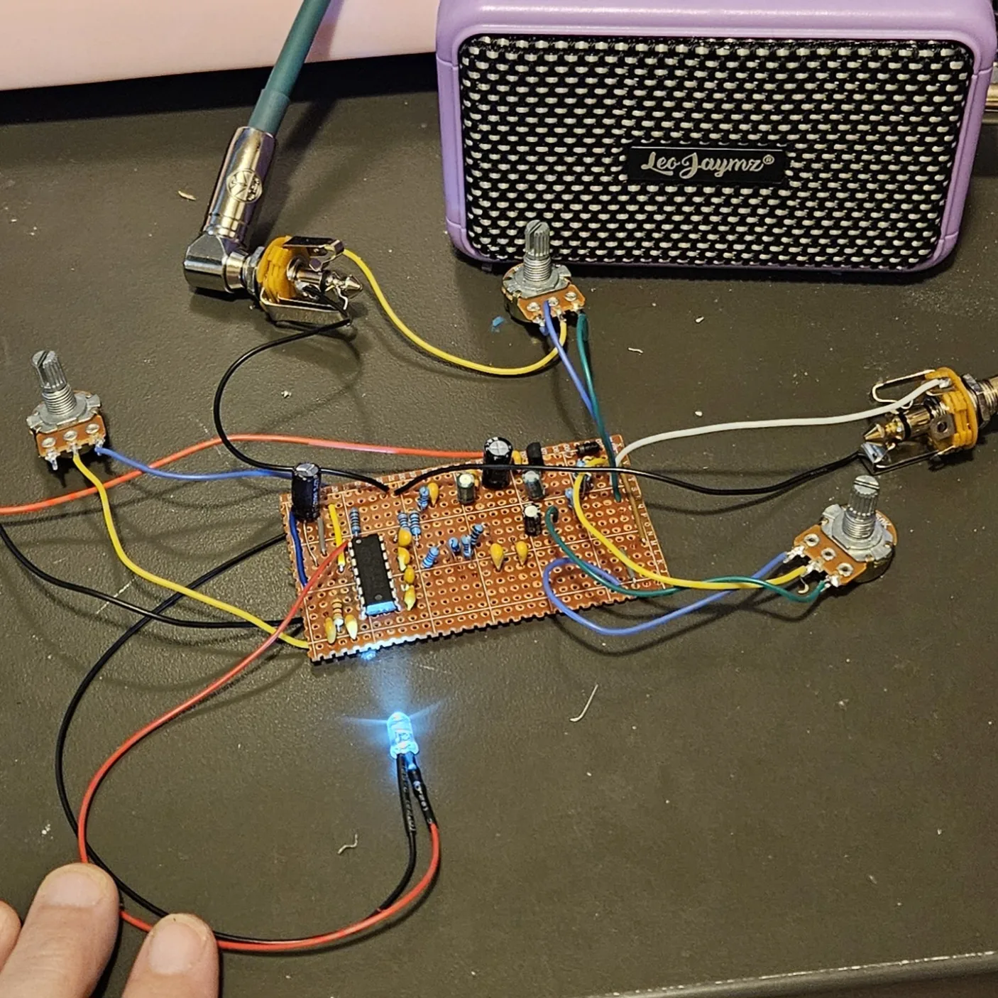 The finished board powered up on a dark workbench. A bare stripboard carries one integrated circuit at its left, rows of upright blue-bodied resistors, orange and tan capacitors and several black electrolytics. Wires in red, black, yellow, blue, green and white run off every edge to three potentiometers and two jacks lying loose on the bench, one of them with a right-angle guitar cable plugged into it. A lit LED on two flying leads sits on the bench in front of the board. A hand at the bottom left holds the red and black supply leads. A small purple practice amplifier stands behind the board.