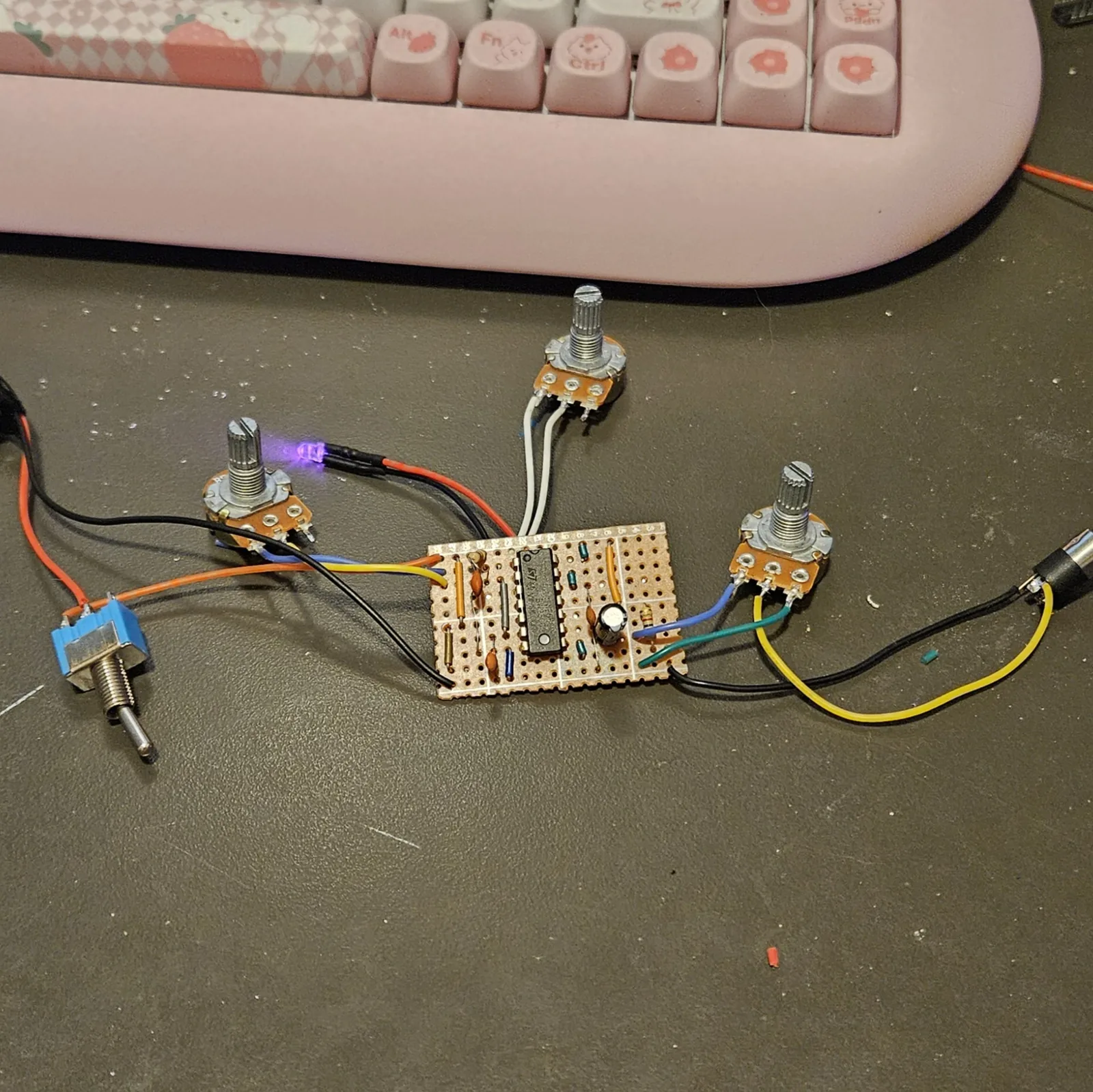 The built board on a dark desk in front of a pink keyboard, wired but not boxed. A small rectangle of orange stripboard in the middle carries the NE556, small capacitors, resistors, one electrolytic capacitor and blue link wires. Three pots hang off it on flying leads: one to the left on yellow, blue and black wires, one above on white wires and one to the right on blue, green and yellow wires. A blue toggle switch lies at the lower left on orange and black leads. A lit LED on red and black leads sits beside the left pot. A black jack lies at the right edge on a yellow and a black wire.