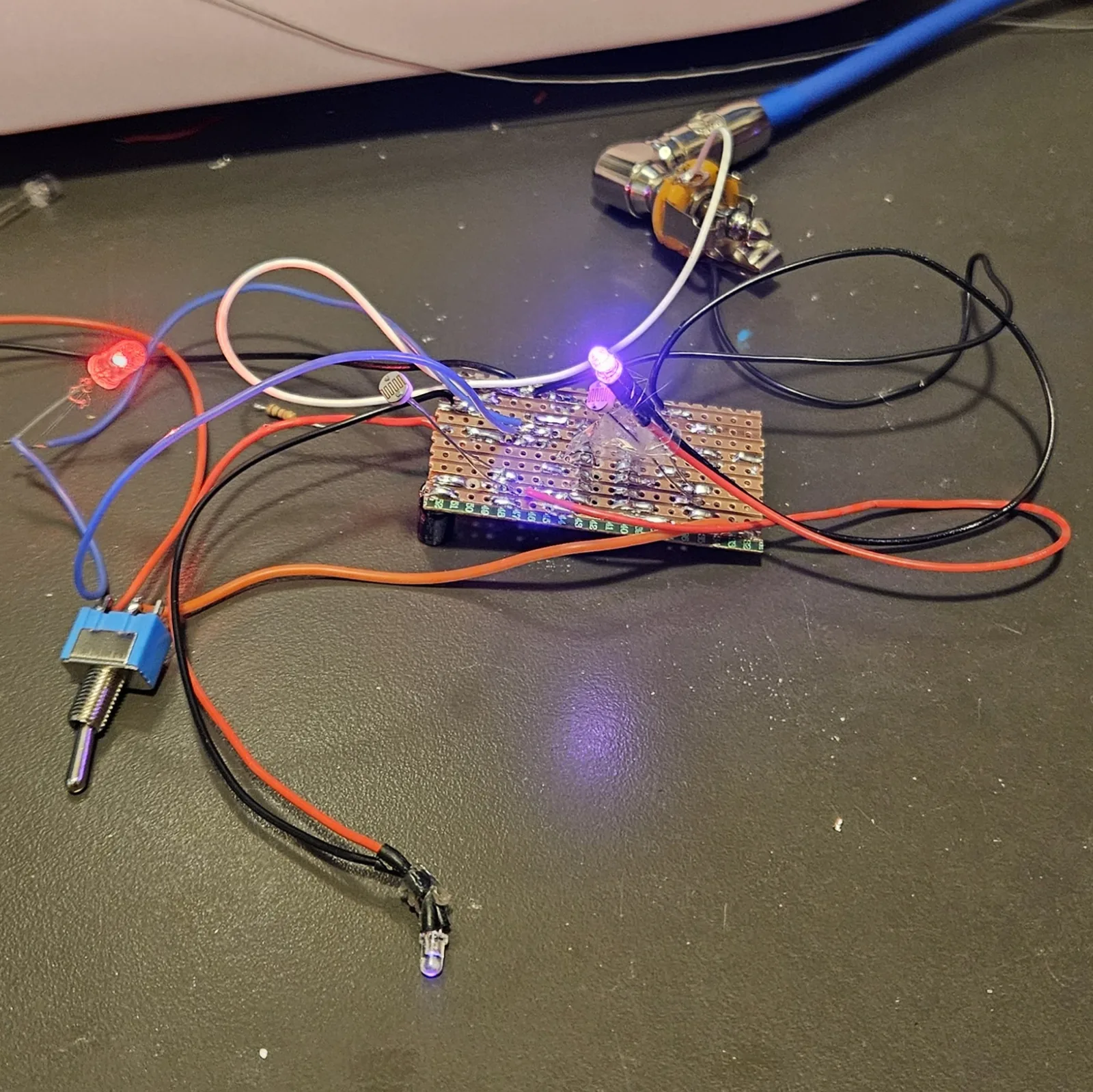 The finished board on a dark desk, working, with everything off the board on flying leads. A small stripboard sits in the middle with its solder side up, rows of bright solder joints along the copper strips. A violet LED on the board is lit, glowing against a photocell fixed beside it in a blob of clear glue; a second photocell stands at the left edge of the board on white wires. A red LED glows at the far left on red leads, a second violet LED lies at the bottom on black and red leads, and a blue toggle switch lies at the lower left on red and orange wires. A quarter-inch jack at the top right has a blue instrument cable plugged into it. Red, black, blue, white and orange wires loop across the desk between them.