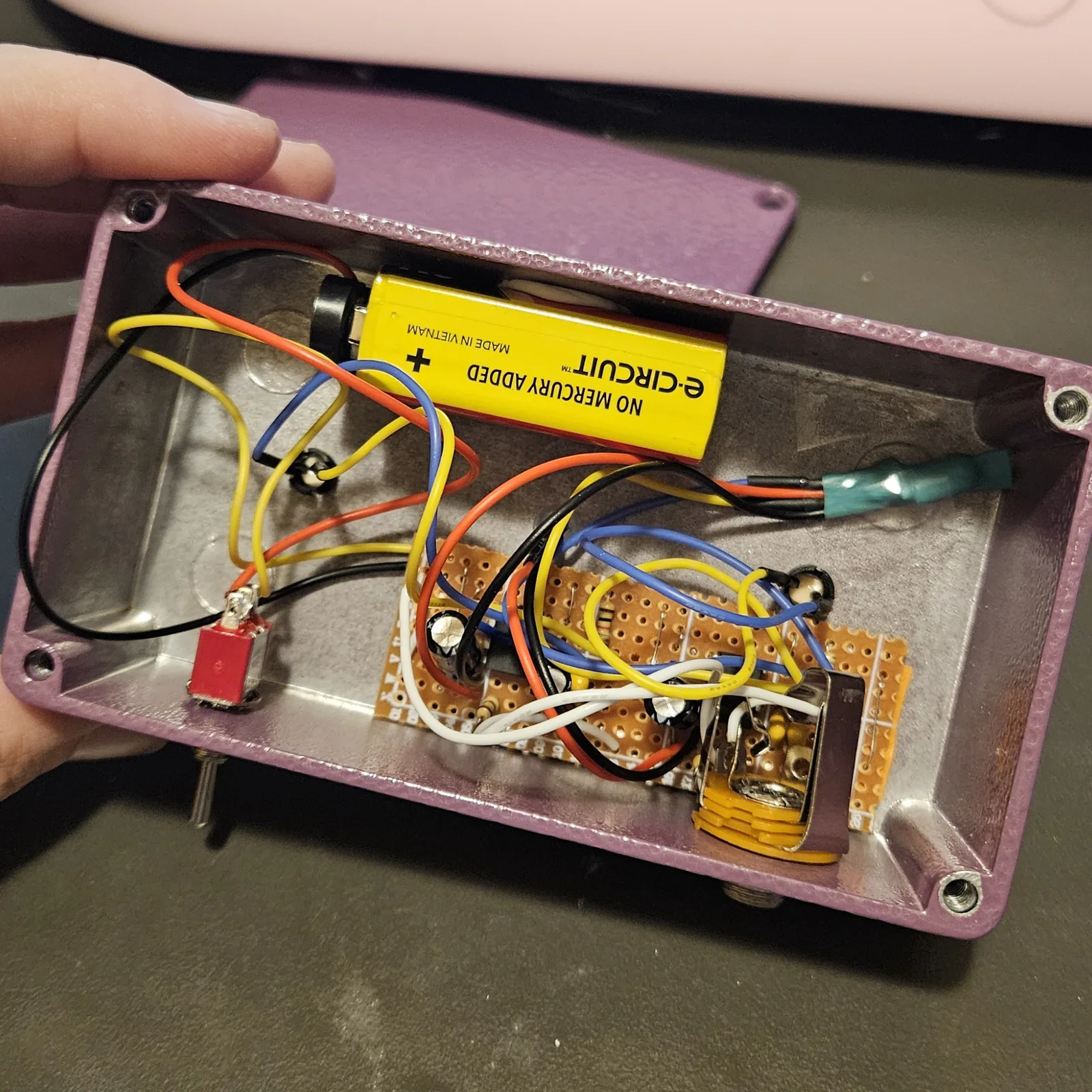 Looking down into the open enclosure with everything inside: a yellow 9-volt battery lying across the top, a red toggle switch mounted through the left wall, the stripboard flat in the middle with its electrolytic capacitors standing up off it, the quarter-inch jack at the lower right, and orange, black, blue, yellow and white wires running between them. A teal heat-shrink sleeve sits along the right-hand wall.