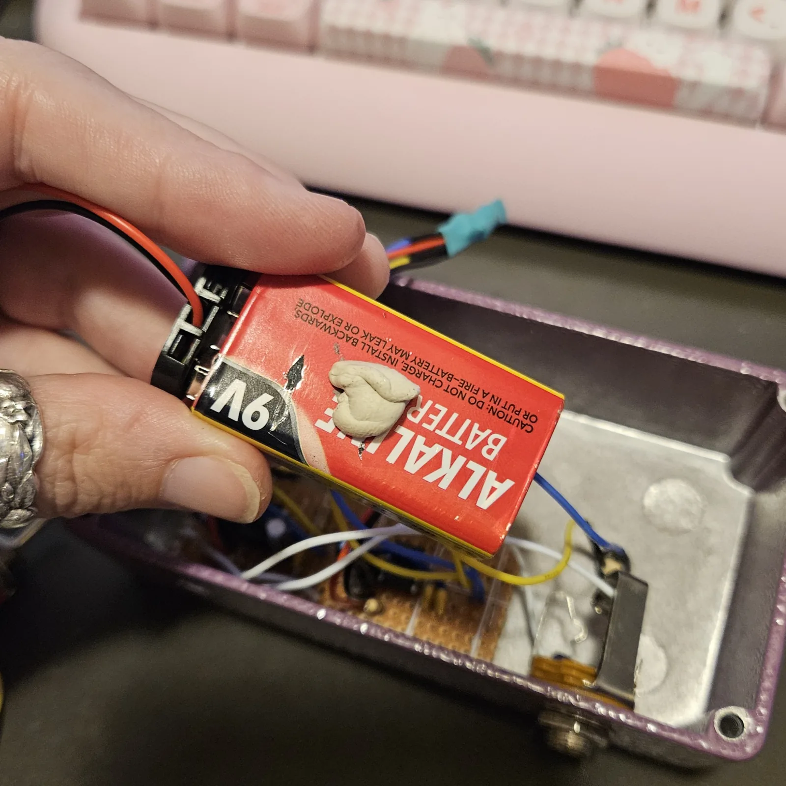A red 9-volt alkaline battery on a black snap connector, with a blob of white putty stuck to its side, held above the open enclosure and about to be pressed down on top of the wired board.
