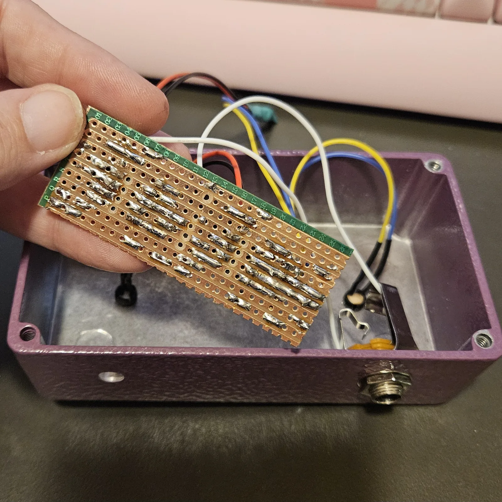 The finished stripboard held over the open enclosure, solder side toward the camera: rows of holes joined by bright blobs of solder along nine copper strips, with the column numbers printed down the green edge of the board. Below it the open purple enclosure already has its toggle switch and quarter-inch jack mounted, with orange, black, blue, yellow and white wires looping out of them.