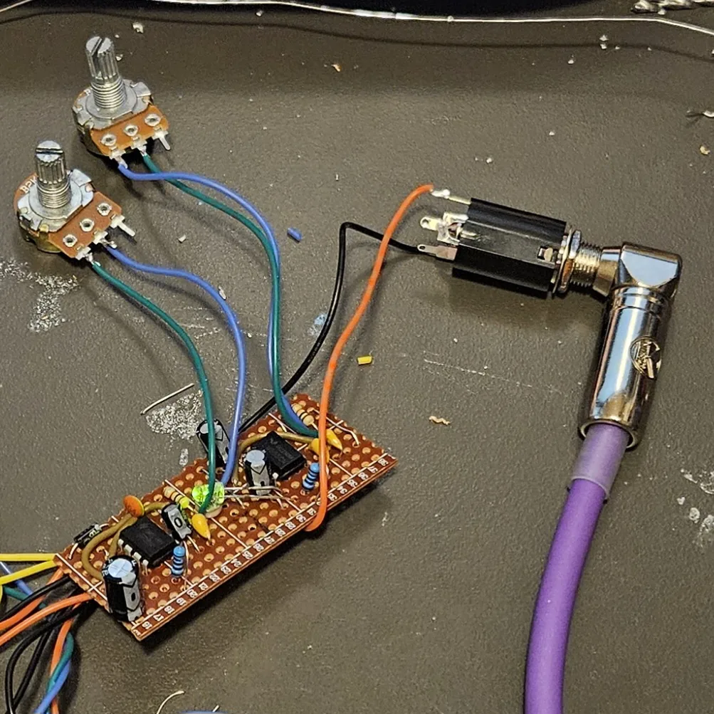 The Opto Duet v2 board on a dark workbench mat, not yet boxed. A narrow strip of brown stripboard at the lower left carries two black eight-pin chips, small blue resistors, yellow ceramic capacitors, black electrolytic cans and a small green LED, with a bundle of yellow, orange, black and blue wires leaving its left end. Two potentiometers with bare knurled shafts sit above it on green and blue wires, and at the right a quarter-inch jack is wired on orange and black leads, with a purple cable plugged into it through a chrome right-angle plug.