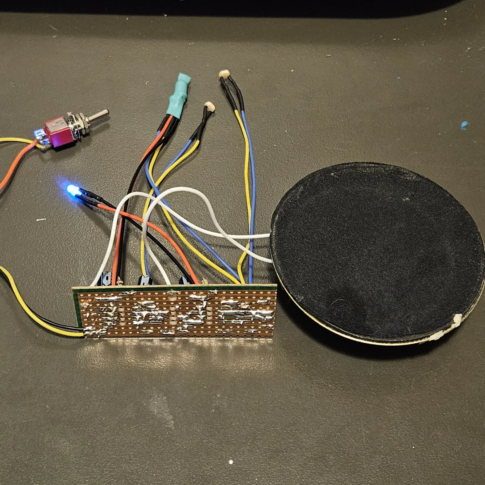 The finished board on a dark desk mat, photographed from the solder side: a long narrow strip of copper-clad board, its rows of holes joined by bright blobs of solder, with flying leads in orange, black, blue, yellow and white leaving the top edge. A red mini toggle switch sits at the upper left on yellow and orange wires, and a blue LED burns bright next to it. A teal heat-shrink sleeve swallows one pair of leads, and two small cream photocells stand on their own black wires. A large black round speaker lies to the right of the board.