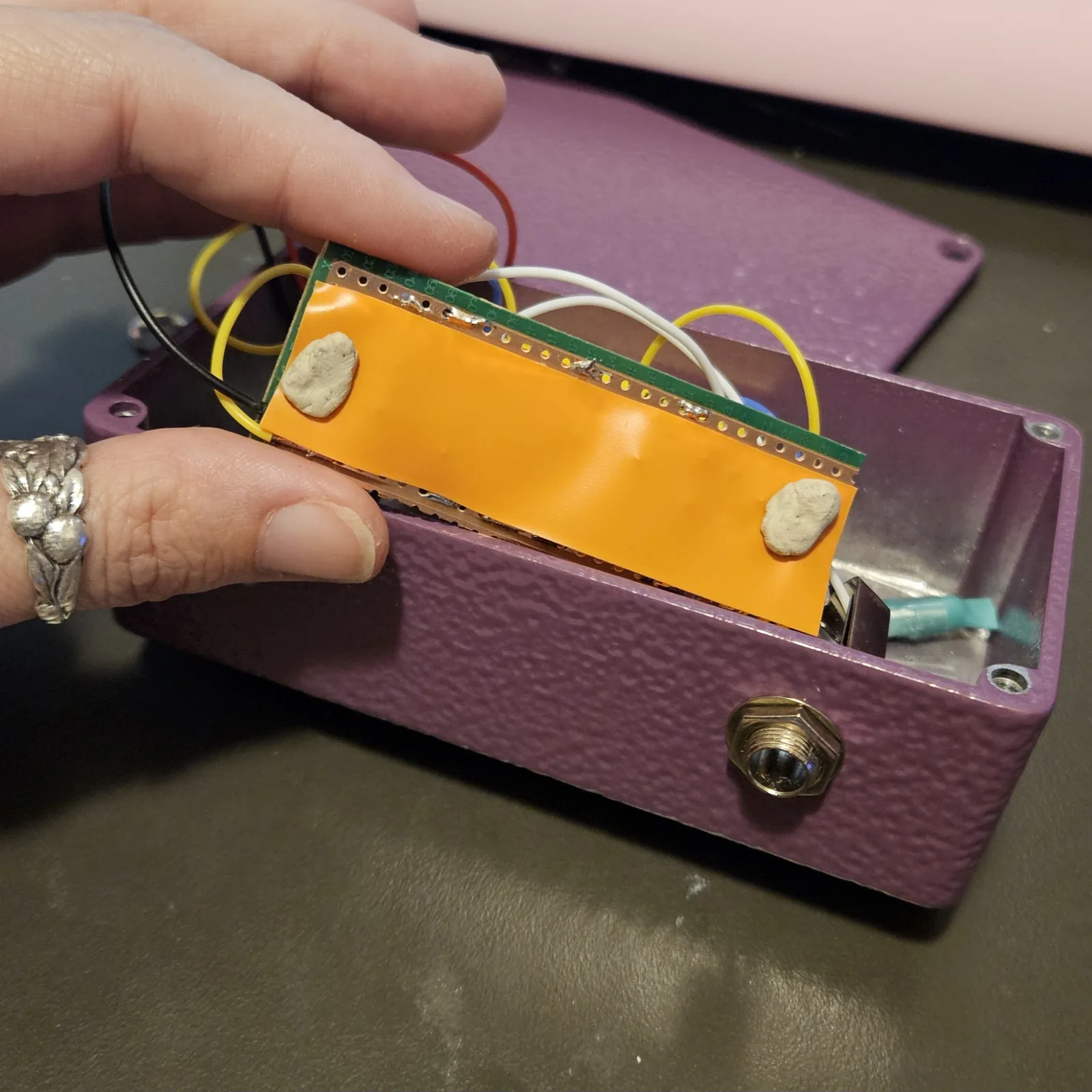 The board going into the enclosure with a strip of orange insulating tape stuck across its whole solder side and two blobs of white mounting putty pressed onto the tape. The purple box below already holds the jack and the switch, and a teal heat-shrink sleeve lies inside it.