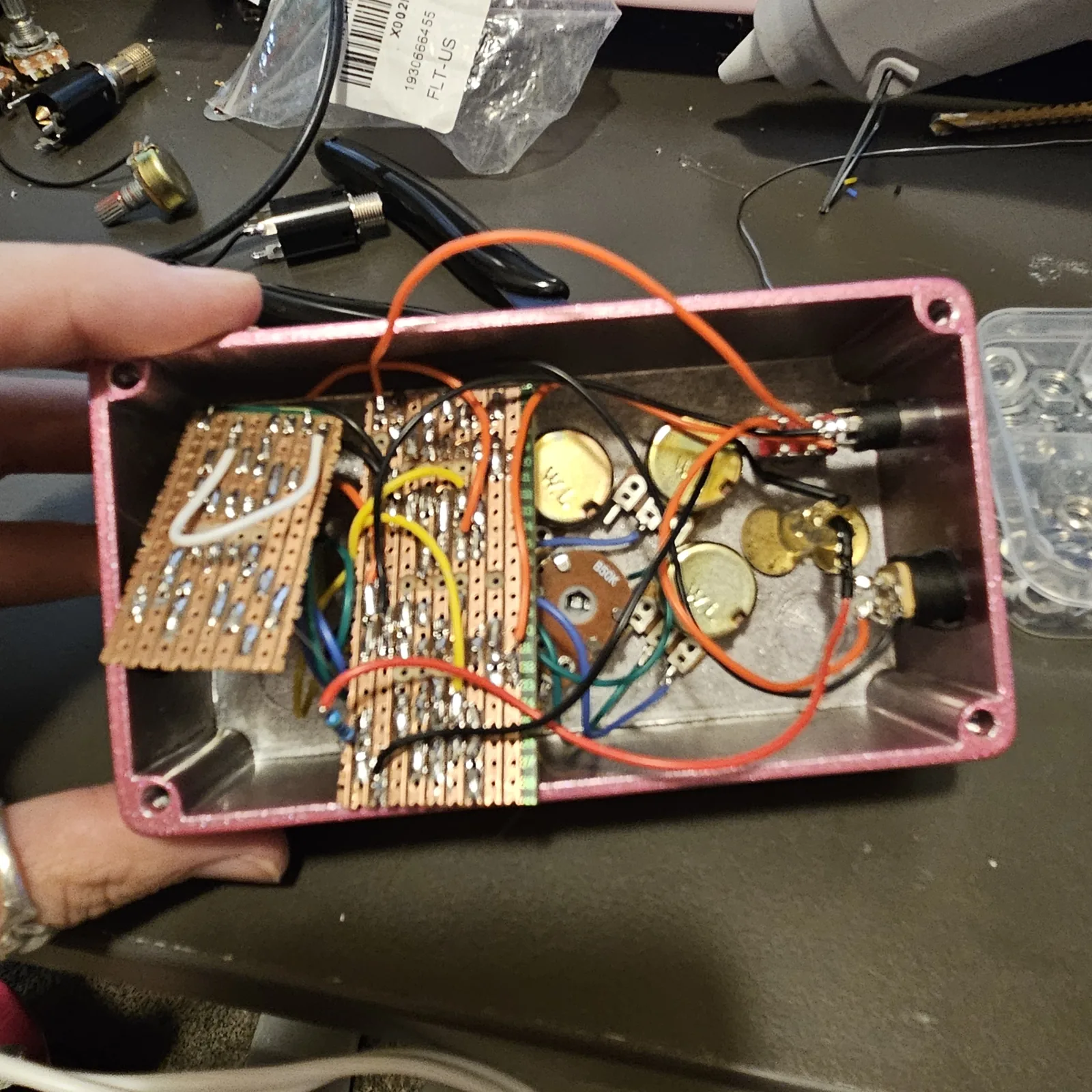 The Opto Wobble opened up and held in one hand over a workbench: a glittery pink 125B enclosure lying on its back with two pieces of stripboard mounted inside, one at the left carrying a row of small resistors and a white wire link, one in the middle crowded with parts, and bundles of orange, black, yellow, blue and green wires running from them to the round metal backs of the pots and to the jacks at the far end. Pliers, a glue gun, a spare jack and a pot lie on the bench around it.