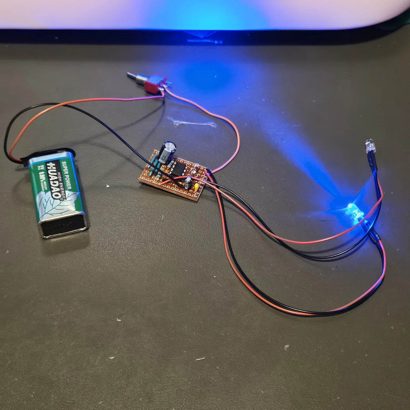 The Police Lights built on a small piece of stripboard, lying on a dark bench with wires trailing off it. The 555 sits in the middle of the board with a black electrolytic capacitor standing beside it and a small yellow capacitor near its edge. To the left a 9 V battery lies on its side, its snap leads running up to a small red toggle switch at the top of the picture. To the right two LEDs hang on long red and black leads; one of them is lit and throws a bright glow across the bench.