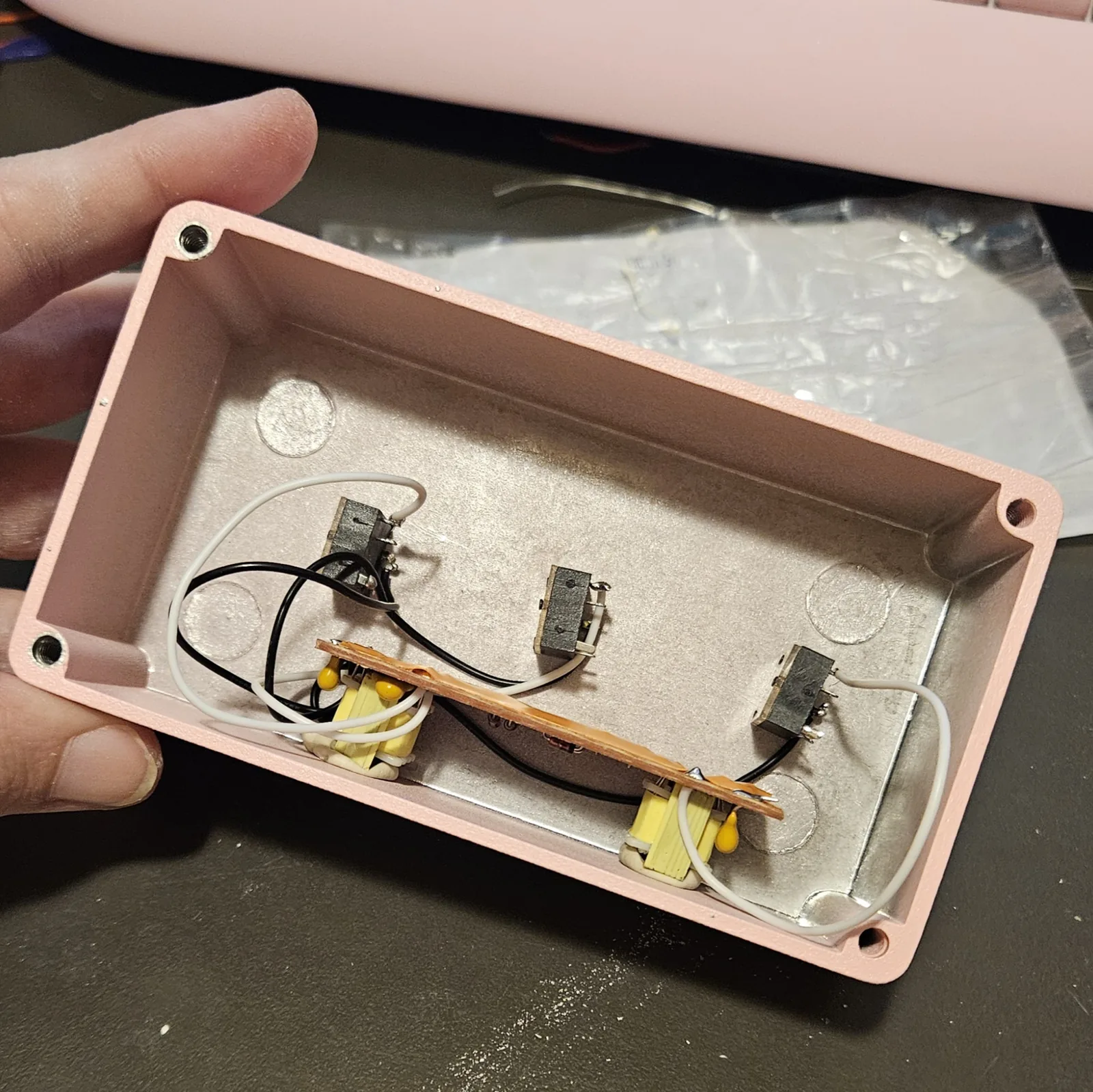 The board lying inside the open pink enclosure, parts down and taped side up, resting on the two putty-wrapped transformers. Three black jacks along the far wall are wired to it with black and white wires. A hand holds the box and a pink keyboard sits behind it.
