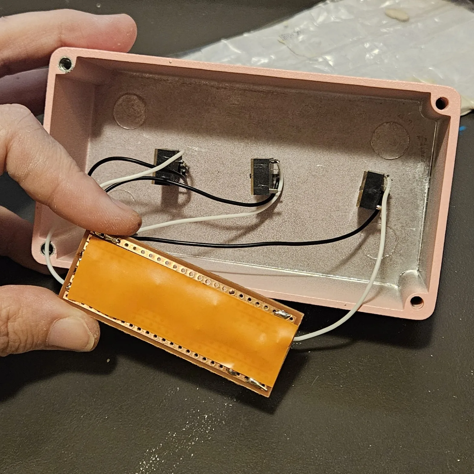 The board turned over to show its solder side, covered edge to edge with a strip of orange insulating tape, held over the open pink enclosure. Three black jacks are mounted along the far wall of the box, with black and white wires running from them toward the board.
