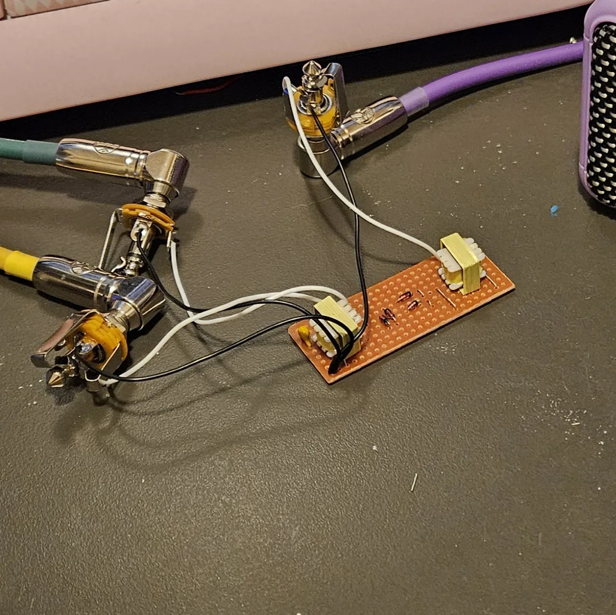 The finished board on a dark desk mat, wired but not boxed. A short strip of orange-brown stripboard lies at the lower right with a small cream-and-yellow transformer at each end and a few tiny glass diodes and wire links between them. Black and white wires run off its left end to three quarter-inch jacks lying loose on the mat, two together at the left and one at the top; the top jack has a purple instrument cable plugged into it that runs off toward the corner of a small lilac practice amp at the top right. The edge of a pink keyboard shows along the top of the frame.