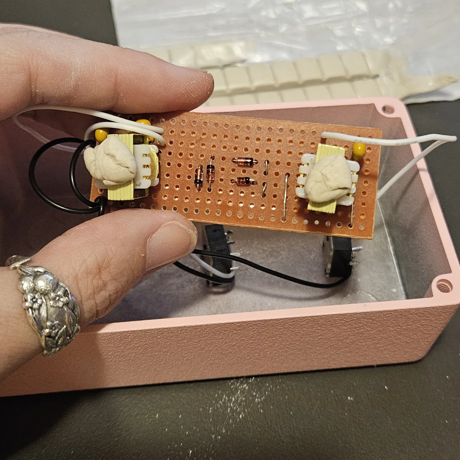 The board held in a hand over an open pink metal enclosure, component side up. Each of the two transformers at the ends of the board is wrapped in a lump of beige mounting putty. Between them are four small glass diodes, two of them standing on end, short wire links, and yellow film capacitors at the corners. Below the board, three black quarter-inch jacks are already mounted along one wall of the box. Behind it a sliced block of white putty sits in a plastic bag.