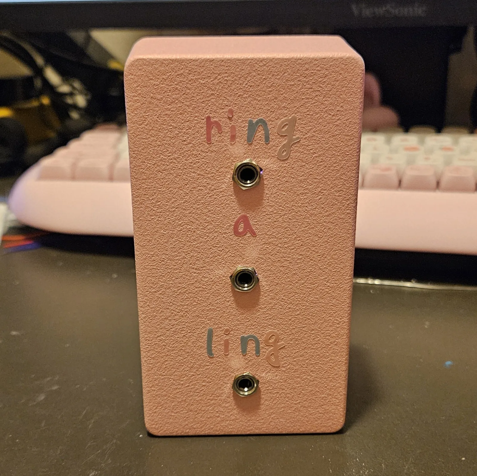 The finished pedal: a pink 125B enclosure standing upright on a dark desk in front of a pink keyboard and a monitor. Three quarter-inch jacks run in a vertical line down the center of the face. Pastel vinyl letters spell ring a ling around them - ring above the top jack, a between the top and middle jacks, ling above the bottom jack - in mixed pink, blue, gray and peach.