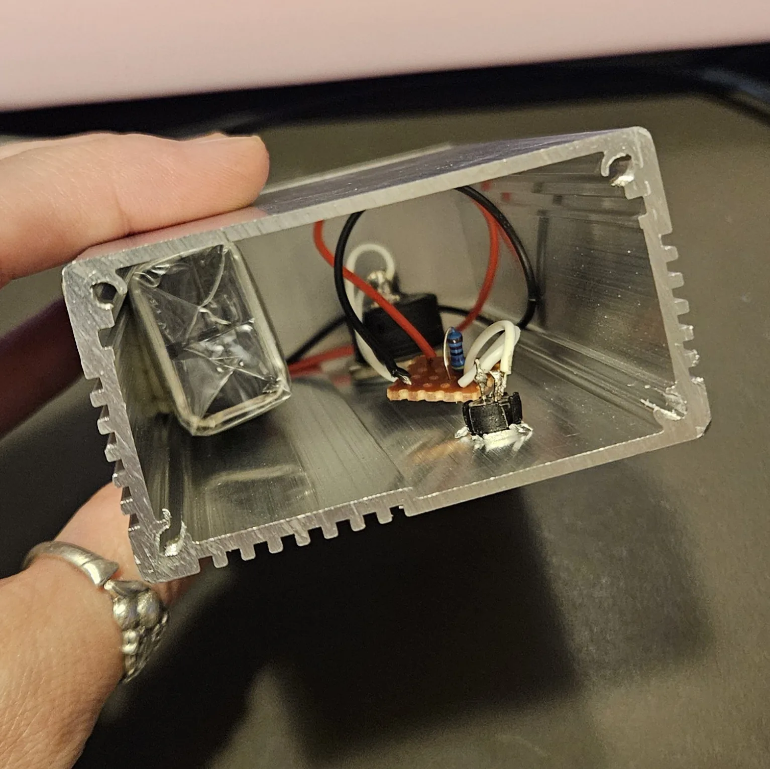 The Button built, seen into the open end of its finned aluminum box, held in a hand. Inside: a 9V battery wrapped in clear tape on the left, the black body of the button with a red and a black lead in the middle, and the tiny orange stripboard beside it with its one blue-bodied resistor standing up. Two white wires run from the board to the back of the LED holder at the bottom right.