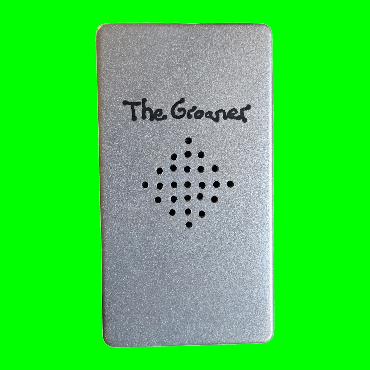 The Groaner - an upright brushed silver box with The Groaner hand-lettered in black across the top and a diamond of small drilled holes below it for the speaker, on a chroma green tile