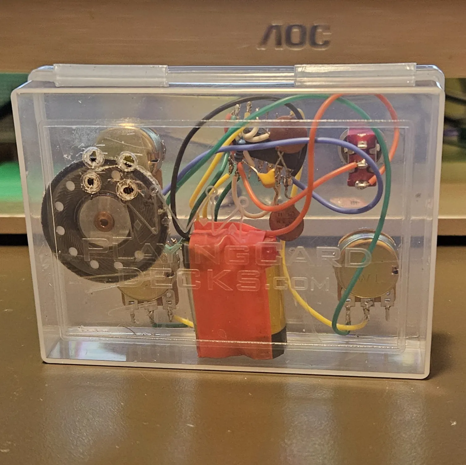 The closed box seen from the back, with PLAYINGCARDDECKS.COM embossed across the clear plastic. The whole circuit shows through it: the black speaker magnet at left, the gold pot bodies, the red toggle, the red-taped battery pack, and the nest of colored wire behind them.