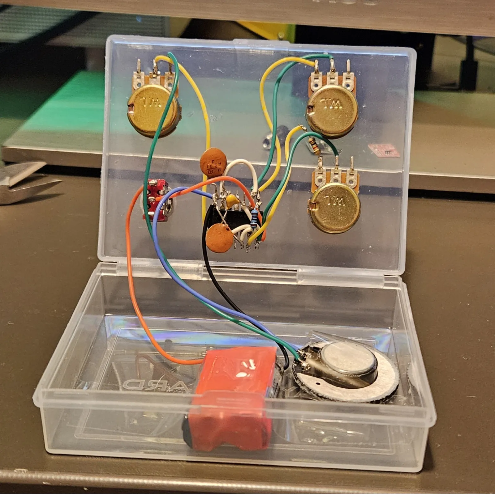 The card box open like a clamshell. Three gold W.L. potentiometers and a red toggle switch are mounted through the lid, with the NE556 soldered dead-bug in the middle of them - legs up, orange ceramic capacitors and a color-banded resistor wired straight to its pins, and yellow, green, orange, blue, white and black hookup wire running out to each control. The lower half of the box holds the red-taped battery pack and the salvaged toy speaker.