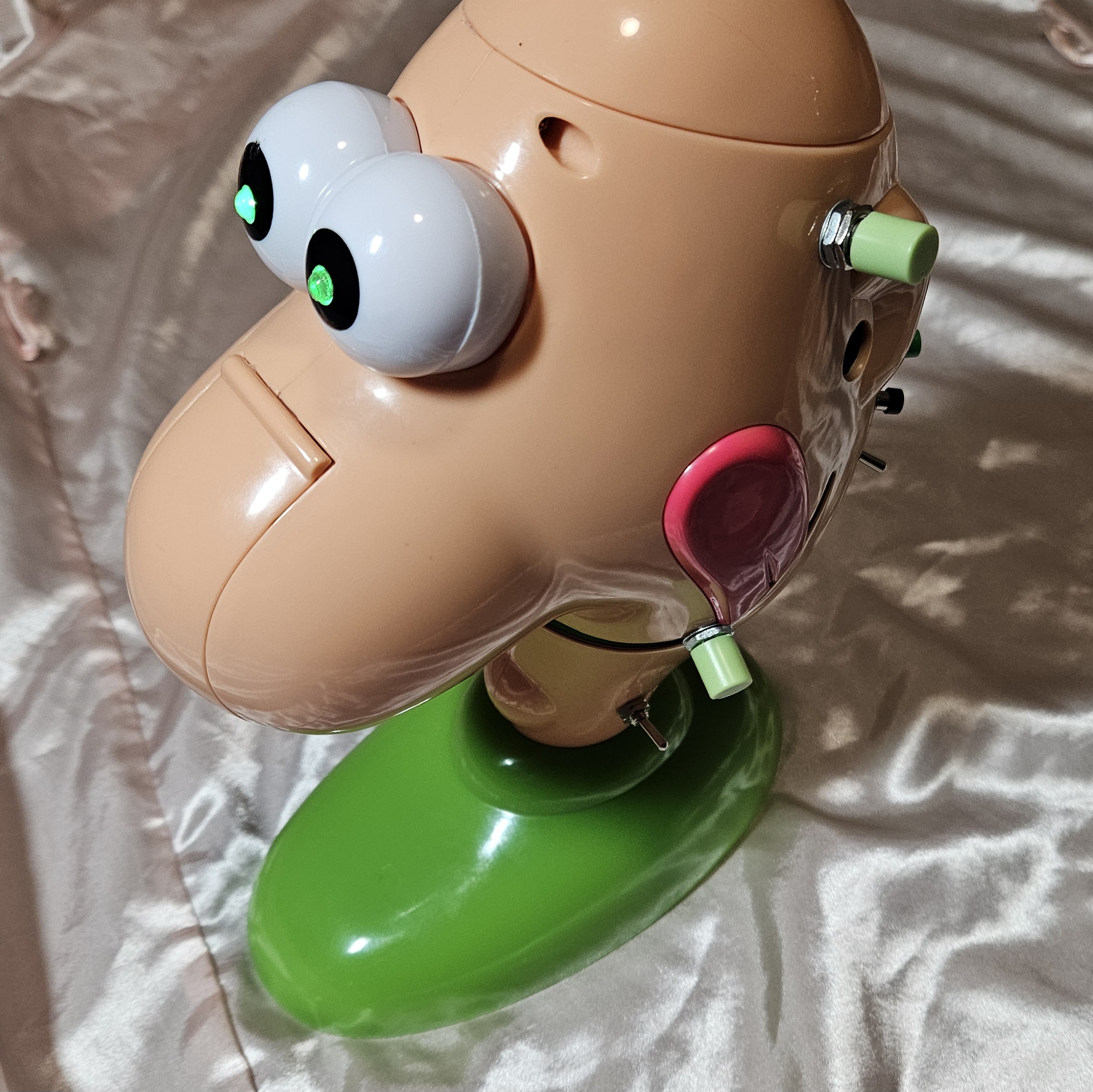 The finished box from above and behind, eyes lit green: the added hardware runs down the right side of the head &mdash; a pale-green cylindrical button cap on a chrome nut high on the back, a smaller green cap and a black cap below it, and a silver mini toggle lever. A second pale-green cap on a chrome nut is mounted low near the tongue, with a chrome toggle just above the green base.