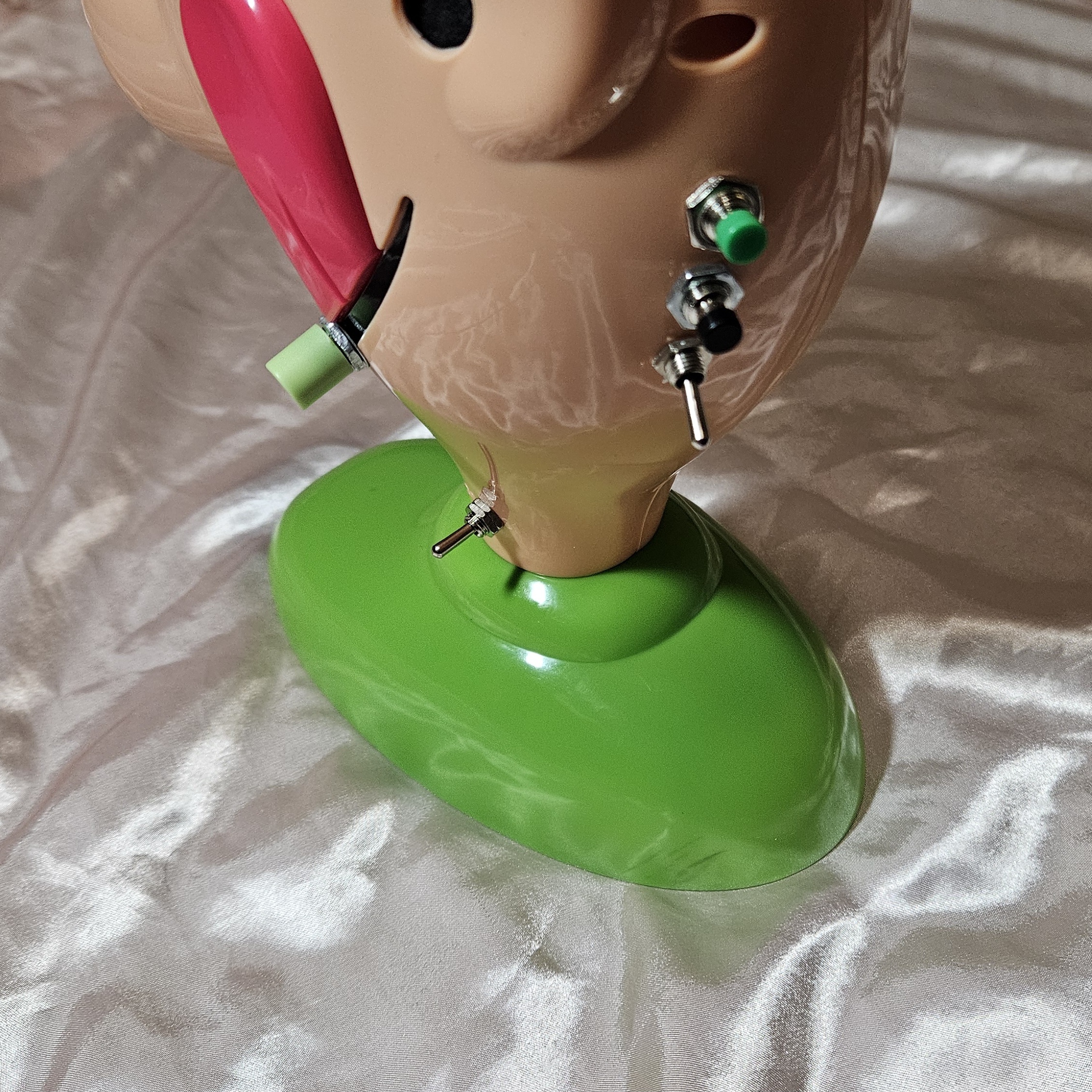 Tight view of the added controls on the head and neck: a green pushbutton cap on a chrome nut, a black pushbutton cap on a chrome nut below it, and a chrome mini toggle with its silver lever pointing down, all in a vertical line on the side of the head. A pale-green cylindrical cap on a chrome nut sits on the far side near the pink tongue, and a second chrome mini toggle is fitted low in the neck just above the green base.