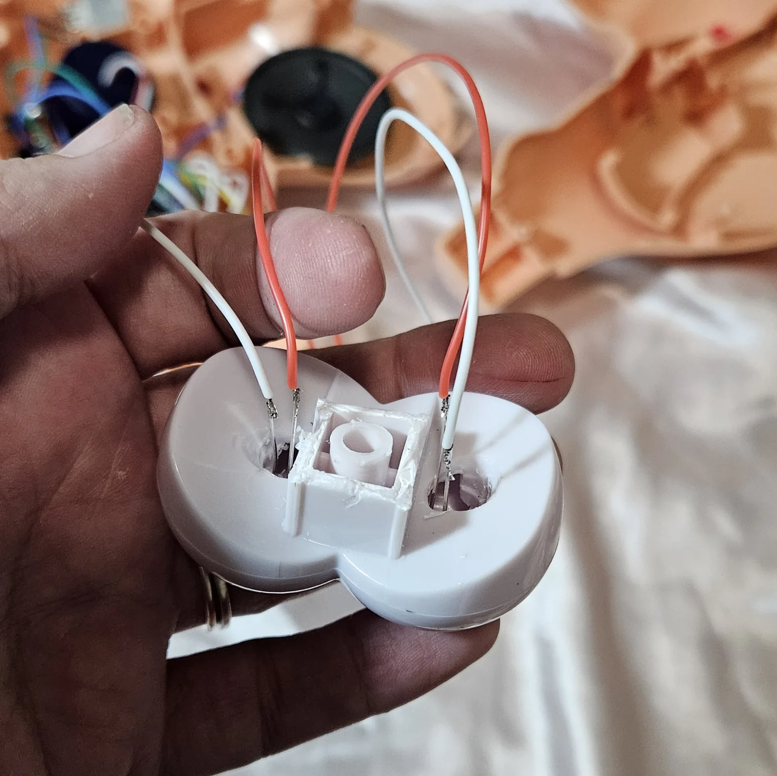 The eyeball assembly held in a hand, back side toward the camera: the white two-lobed plastic piece has a hole drilled through the back of each eye, with component leads pushed through and soldered to a red wire and a white wire on each side. White glue is packed around the central square post. Behind, out of focus, are the opened flesh-colored shell, a black speaker and a tangle of colored wire.