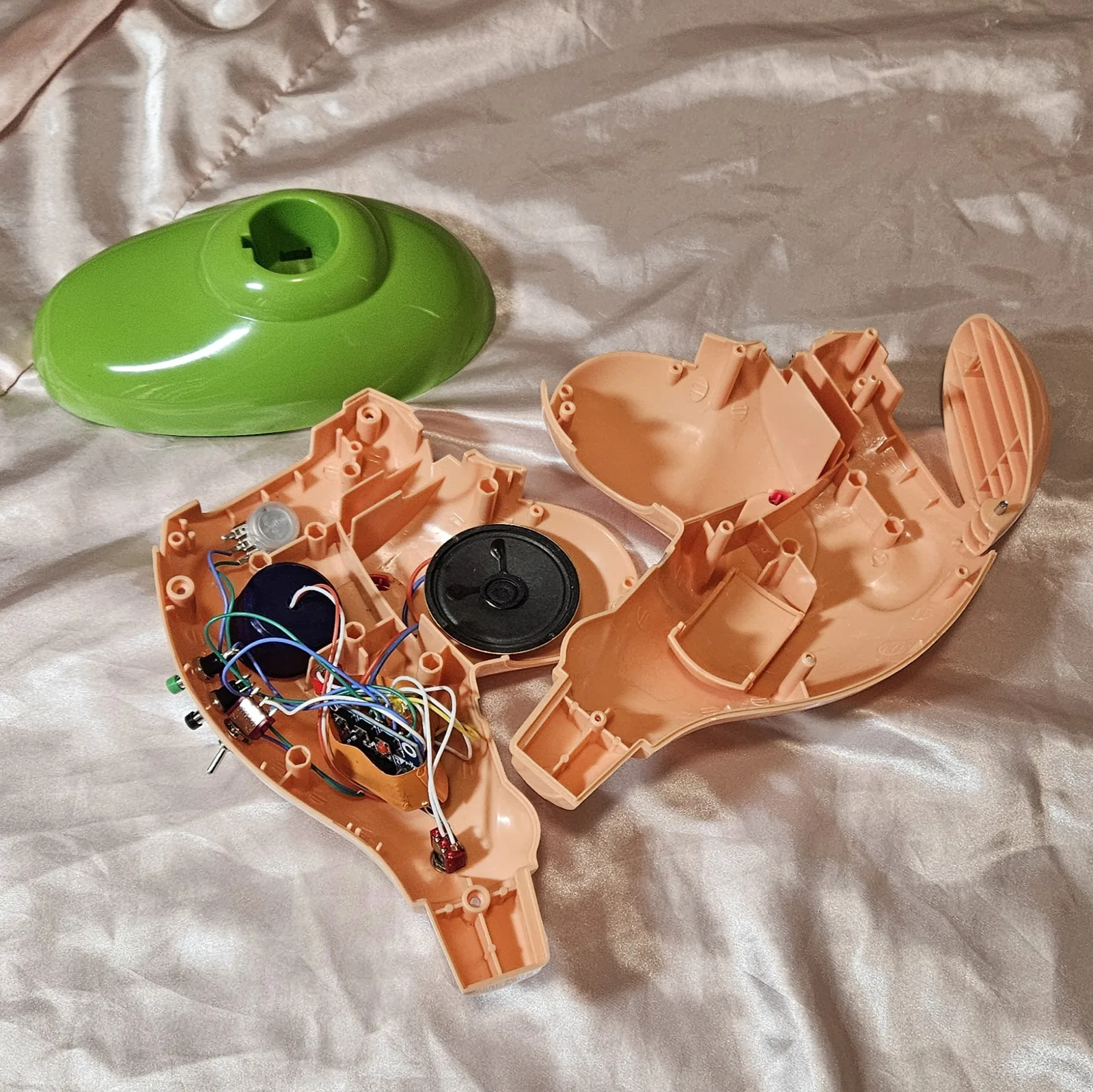 The guts: the left peach half-shell lying open and loaded, with the empty right half and the green base shell beside it. The blue ISD1820 recorder module sits low in the shell, its row of header pins fanned out to colored wire; a round black speaker sits in the middle of the cavity, with a large blue capacitor and a small silver disc toward the top. Green, blue, white, yellow, orange and red hookup wire runs between them in a loose bundle. Along the shell rim are the mounted controls seen from inside - a green pushbutton, a black pushbutton, a silver potentiometer body and two mini toggle switches - and a small red part on twin white wires sits at the bottom.