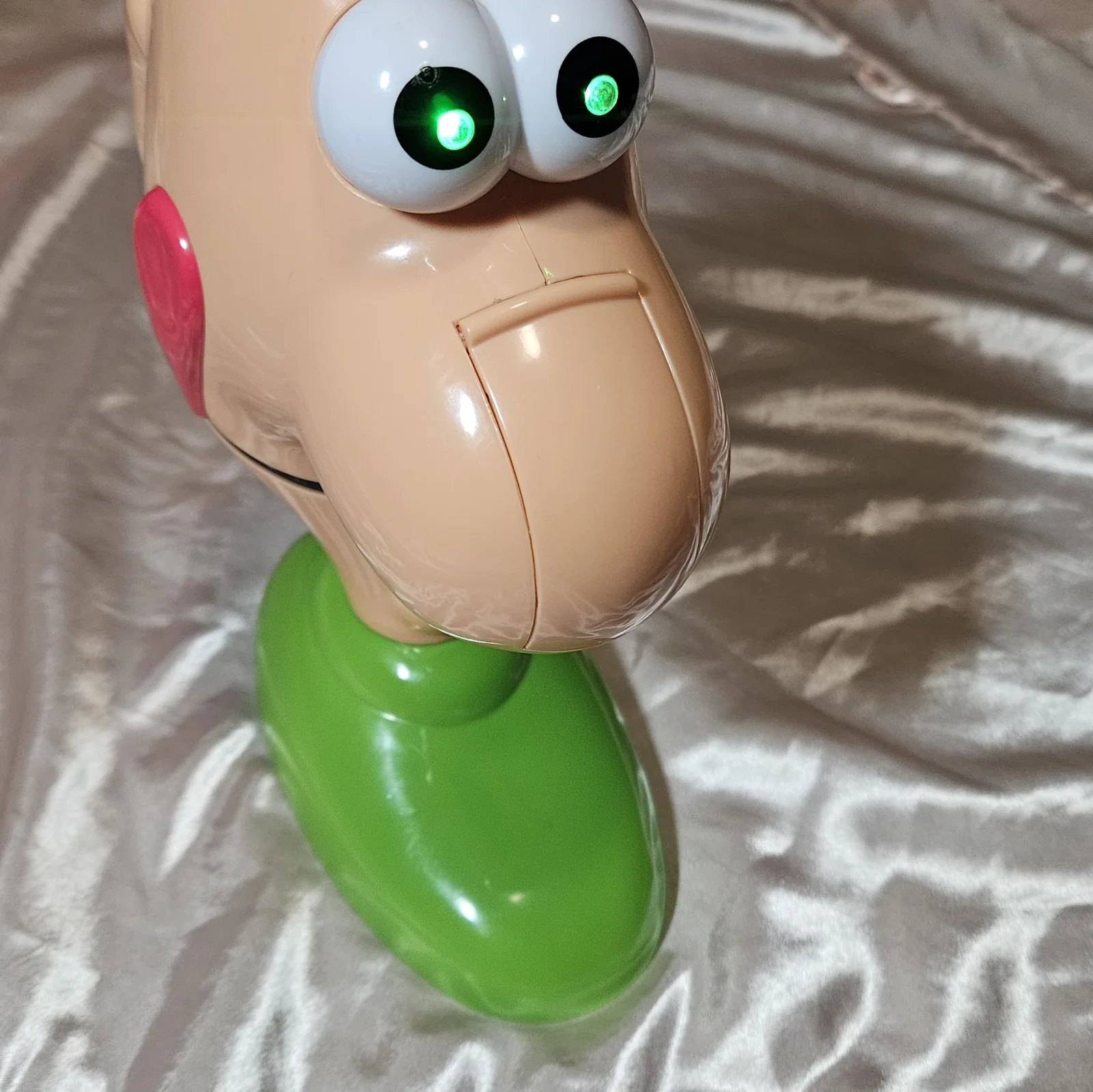 Close-up of the face: both white eyeballs fill the frame with a hard green point of light burning in the center of each black pupil, one flaring brighter than the other. The huge molded nose and the pink tongue at the corner of the mouth are lit only by the room, on shiny cream fabric.