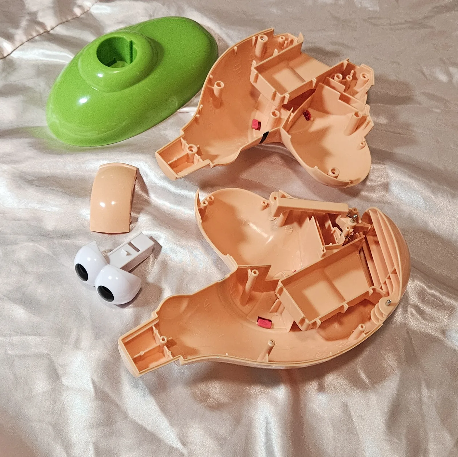 The toy stripped to bare plastic on cream fabric: the hollow green base shell at top left with its keyed socket, the two empty flesh-colored halves of the head laid open to show ribbed molding and small pink plastic tabs, a short curved flesh-colored trim piece, and the white two-lobed eyeball assembly with its black pupils sitting next to a white plastic bracket. No electronics are fitted yet.