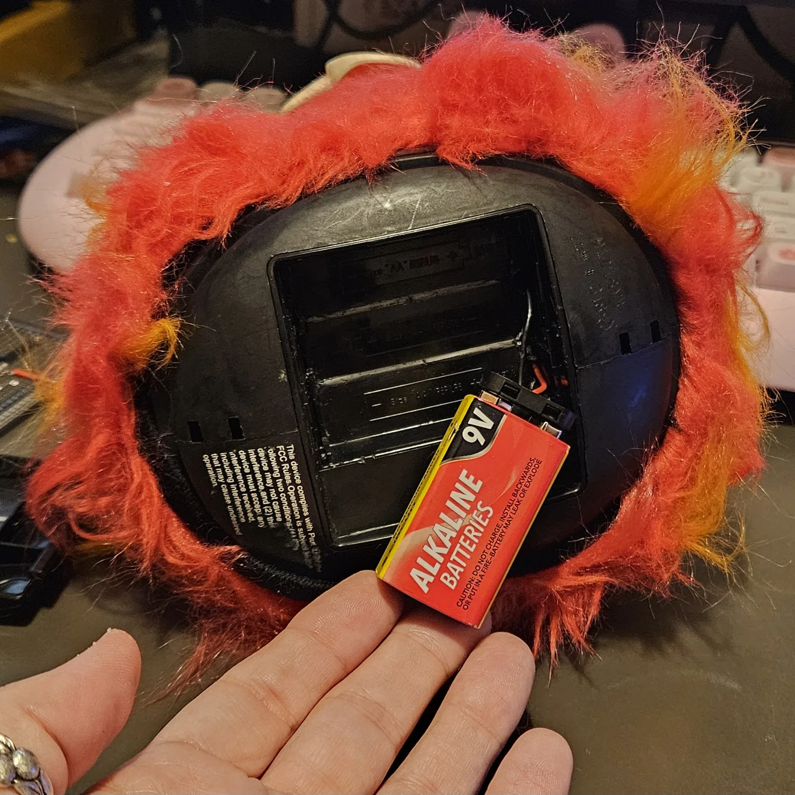 The creature flipped over in one hand: the black oval base with its battery door off, the original AA compartment gutted, and a red 9-volt alkaline battery on a snap connector wedged where the AAs used to live. The molded FCC text is still readable on the shell.
