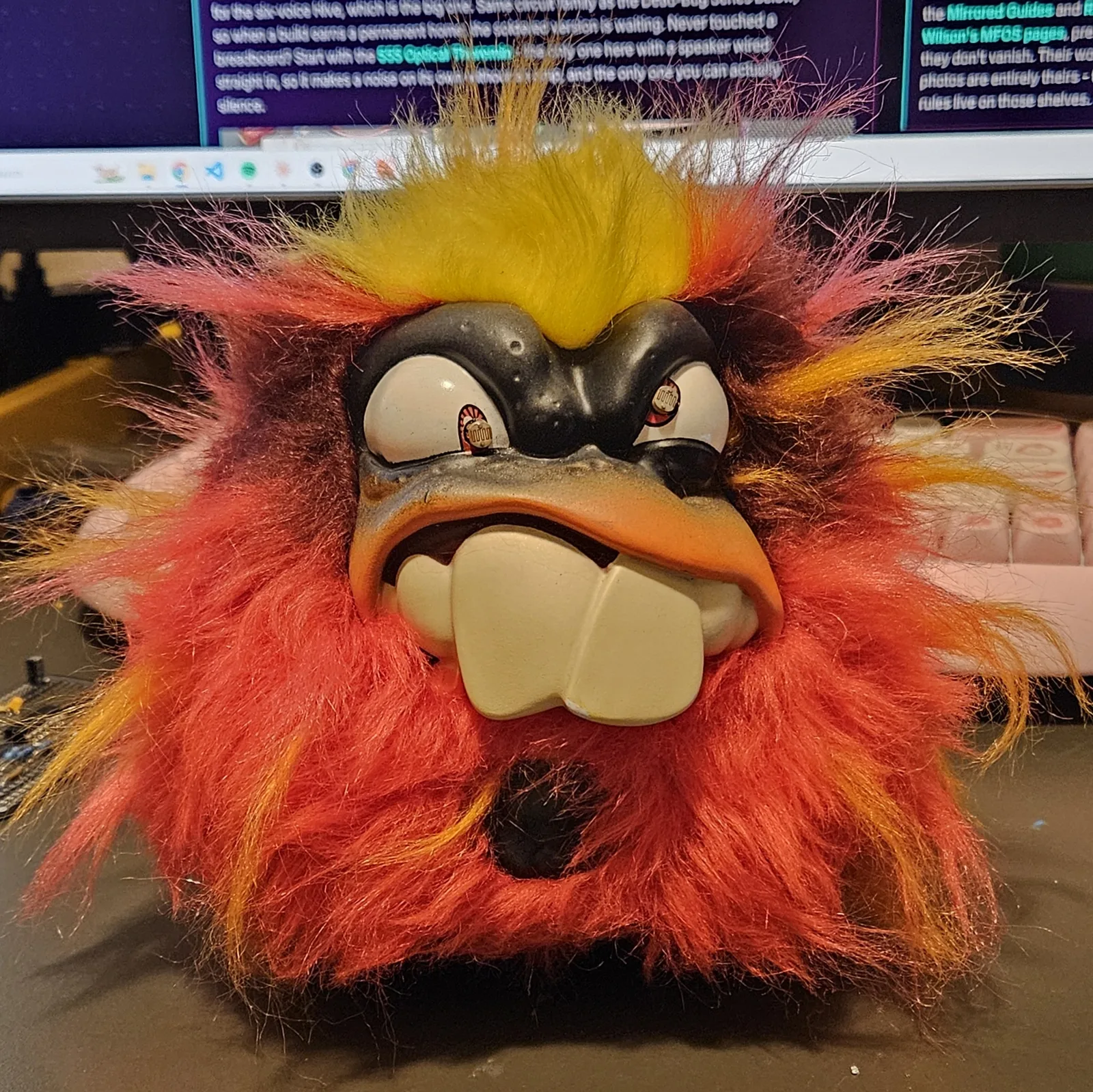 Screaming Creep head on: an angry ball of red, orange and yellow fur with a yellow mohawk, heavy black vinyl brows over wide white eyes, an orange sneering lip and two huge cream buck teeth. In each pupil sits a photocell - a small silver disc with a zigzag track, ringed in orange - staring straight at you.
