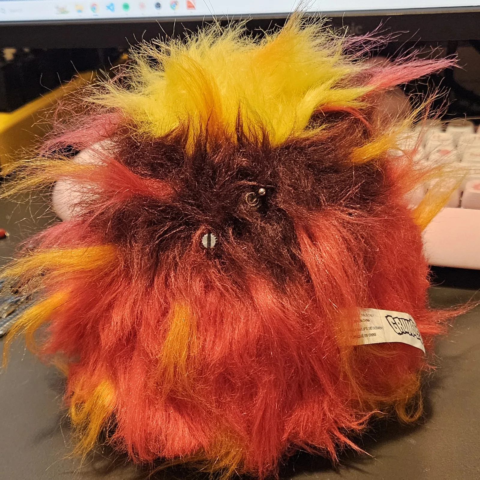 The back of the creature: dark maroon fur parted around two bare metal stems - the slotted volume-pot shaft and the small ball-top on/off toggle - mounted straight through the shell with no knobs, nearly invisible in the fur. The original Grumblies fabric tag pokes out at the lower right.