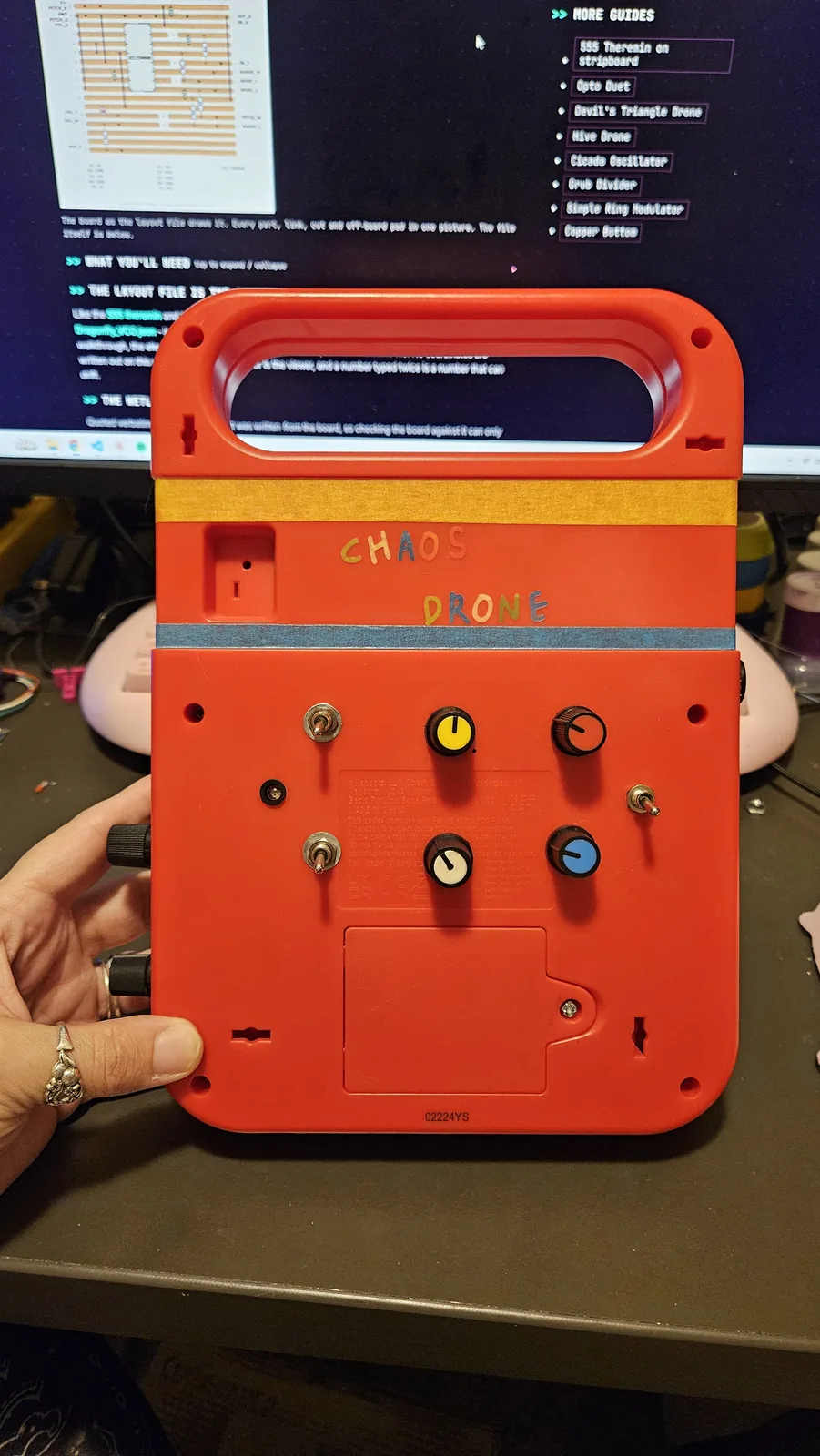 The back of the same Speak and Spell, held in one hand. Two strips of tape run across it, orange near the top and gray below, with CHAOS and DRONE hand-lettered between them in mixed colors. Under the gray strip, seven added controls come straight through the red plastic: four knobs, one yellow, one black, one cream and one blue, arranged in two rows, and three small silver toggle switches around them. The molded battery compartment and its cover are still in place at the bottom.