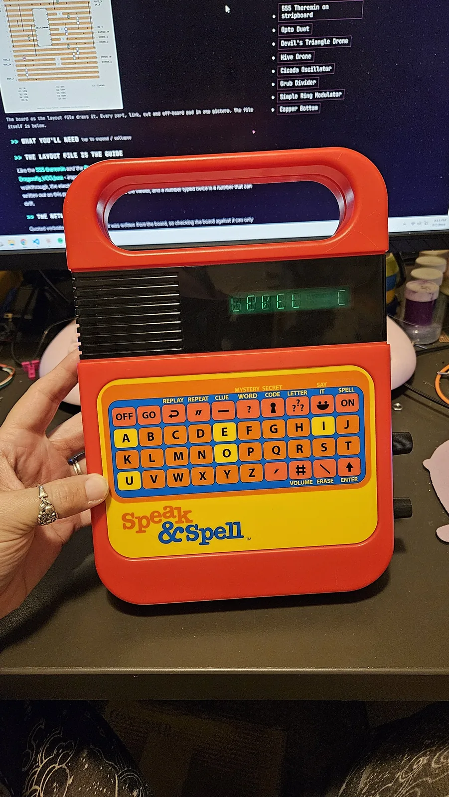 A Texas Instruments Speak and Spell held upright in one hand, face on. The red plastic case has a carrying handle molded across the top and a black panel below it with a row of speaker slots down the left. A green vacuum-fluorescent display in the middle of that panel reads LEVEL C. Under it sits the original keyboard: a yellow panel of small orange letter keys laid out A to Z with a row of blue function keys along the top and down the left, and the Speak & Spell logo in white script at the bottom right.
