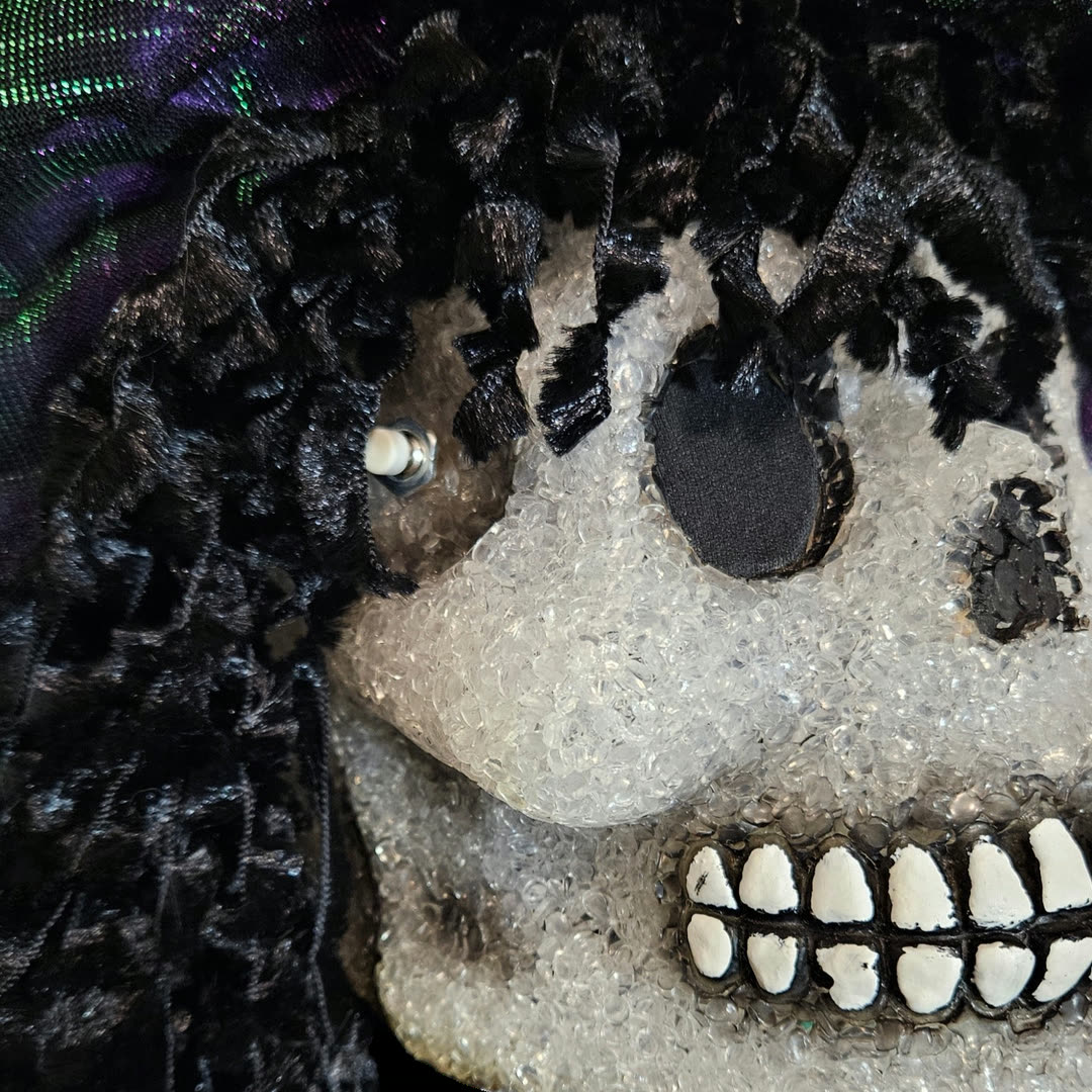 Close-up of the left temple: a small pushbutton with a white tip is mounted through the skull, half-buried in the black crushed-velvet ringlets. Below it the crystalline cheekbone catches the light, and the painted white teeth run along a black-lined jaw.