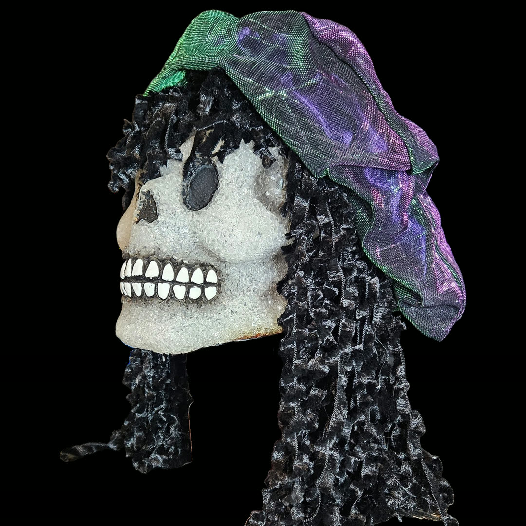 Full left profile against black: the skull's crystalline white face with a black teardrop eye socket and a grinning row of painted white teeth, framed by a long curtain of black ringlets that falls well below the jaw, with the iridescent cap draped over the crown.