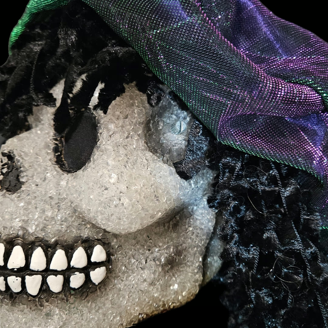 Close-up of the right temple: the pitch-bend potentiometer is mounted straight through the skull with no knob on it &mdash; just the bare metal shaft and its collar, sitting flush in the crystalline finish. The iridescent cap drapes overhead, black curls fall past the jaw, and the painted white teeth run along the lower left of the frame.