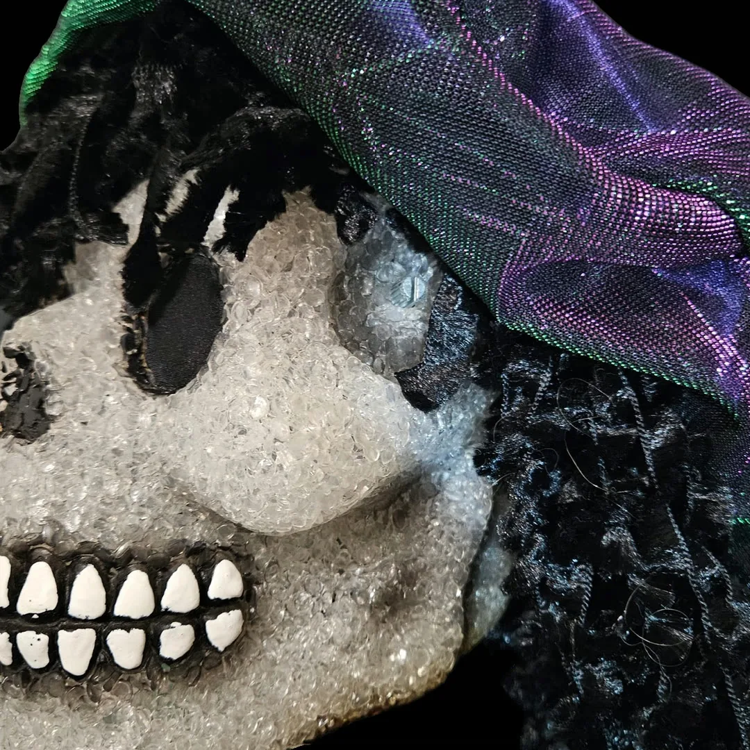 Close-up of the right temple: the pitch-bend potentiometer is mounted straight through the skull with no knob on it - just the bare metal shaft and its collar, sitting flush in the crystalline finish. The iridescent cap drapes overhead, black curls fall past the jaw, and the painted white teeth run along the lower left of the frame.