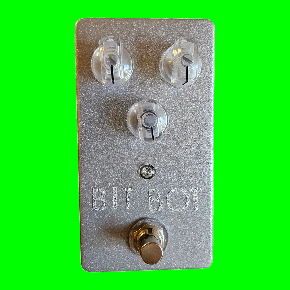 A winked silver pedal photographed face on. Two clear knobs sit side by side near the top with a third clear knob centered below them, a small LED under that, BIT BOT lettered in sparkly silver, and a silver footswitch at the bottom.
