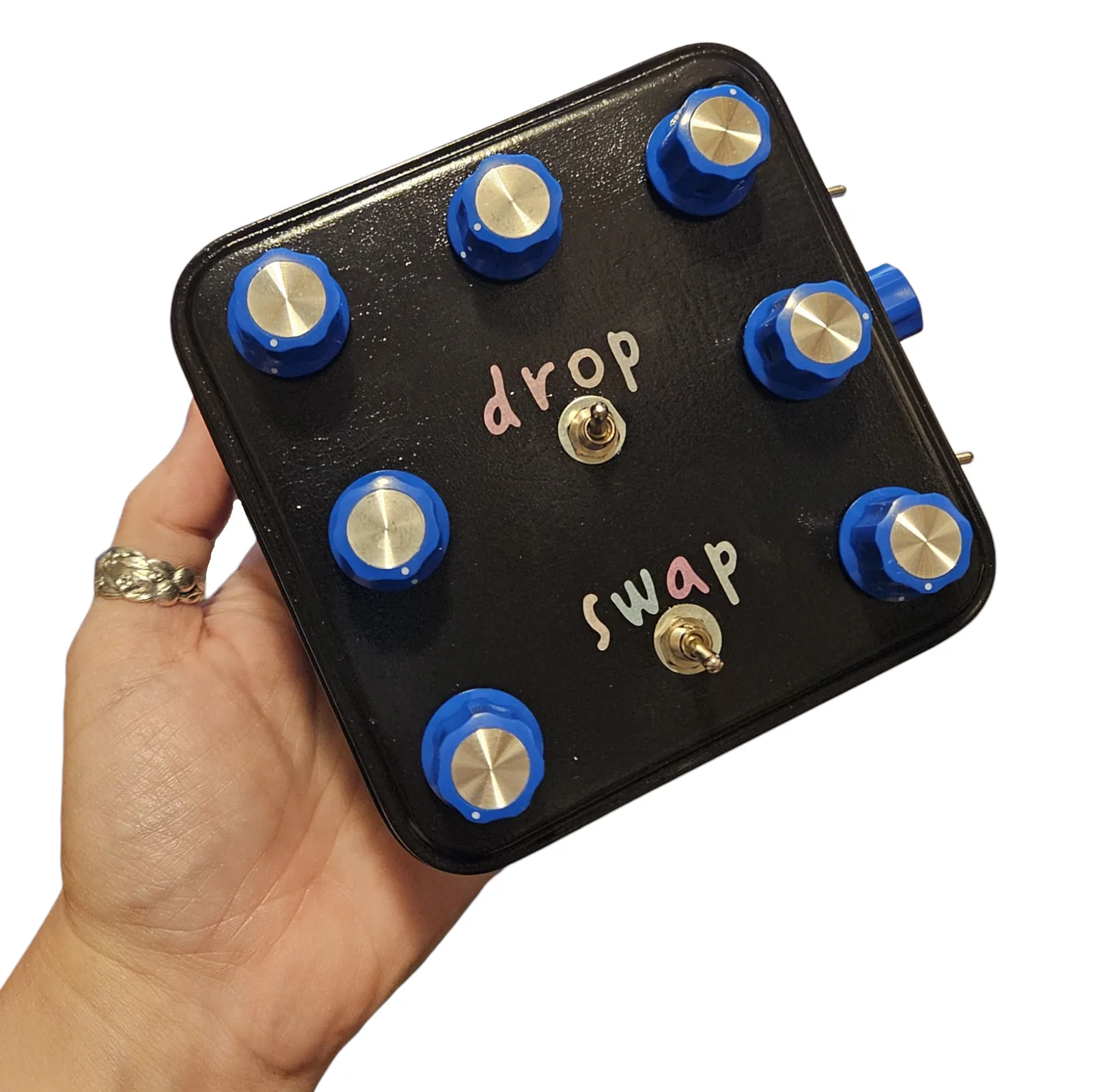 The tin held flat on the open palm of a woman's hand, no bigger than the palm, with a silver ring on her finger. Seven blue knobs around the lid, and the two toggles in the middle labeled drop and swap in small pastel vinyl letters.