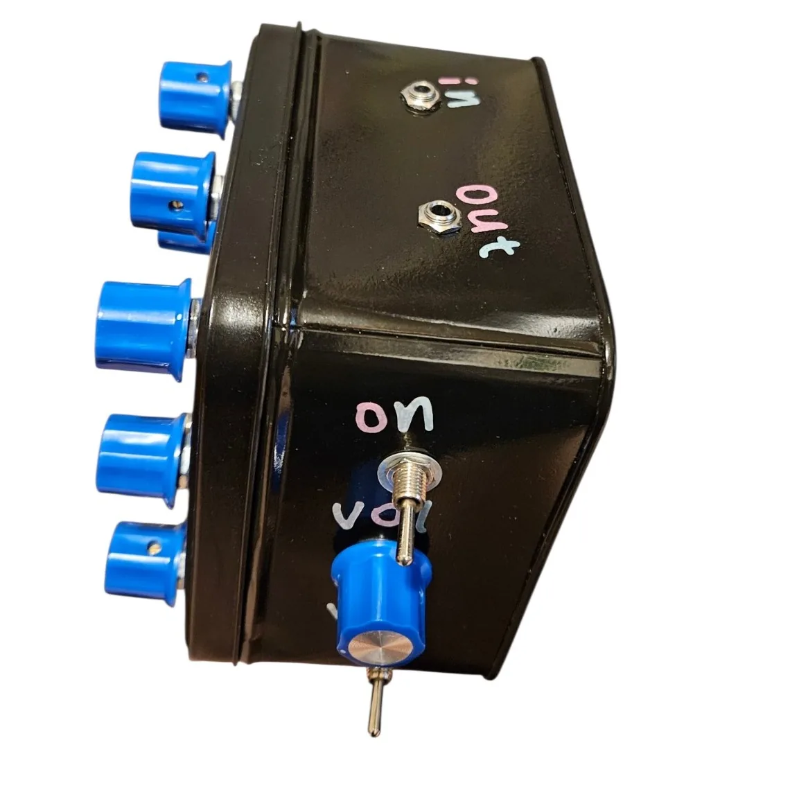 The tin lying on its side against white so its top edge faces the camera: two small 3.5 mm jack sockets set into that edge, labeled in and out in pastel vinyl letters. The lid's blue knobs stand out to the left, and the side panel below shows the on toggle, the vol knob and the vco toggle.