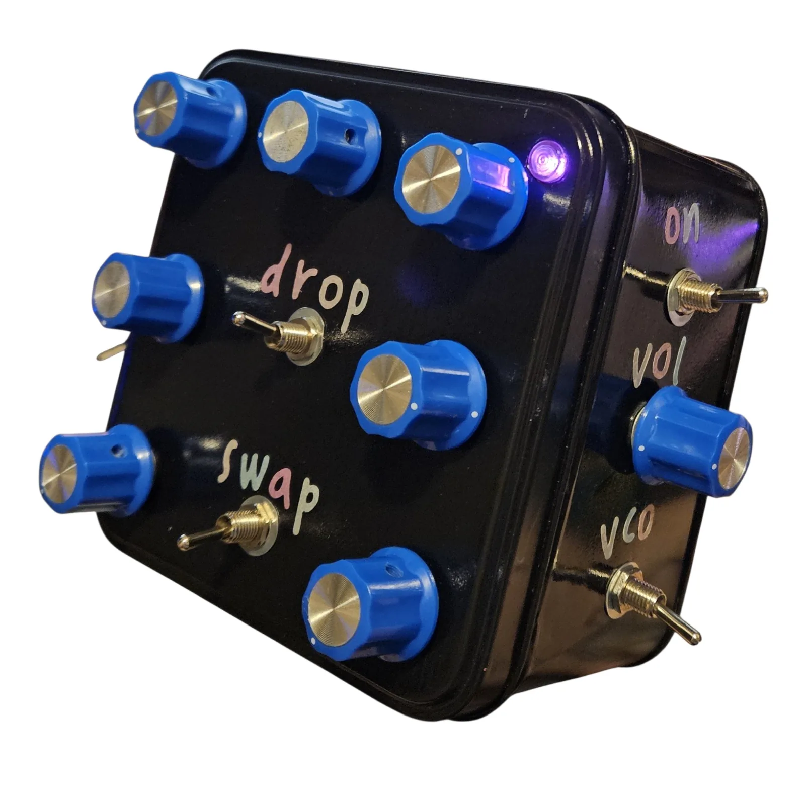 A small square tin painted gloss black, seen from a front three-quarter angle against white with its lid toward the camera and one side showing. Seven blue knobs with silver caps sit around the lid, two chrome toggle switches in the middle are labeled drop and swap in small pastel vinyl letters, and a violet LED is lit in the lid's top right corner. On the side wall a blue knob sits between two toggle switches, labeled on, vol and vco.