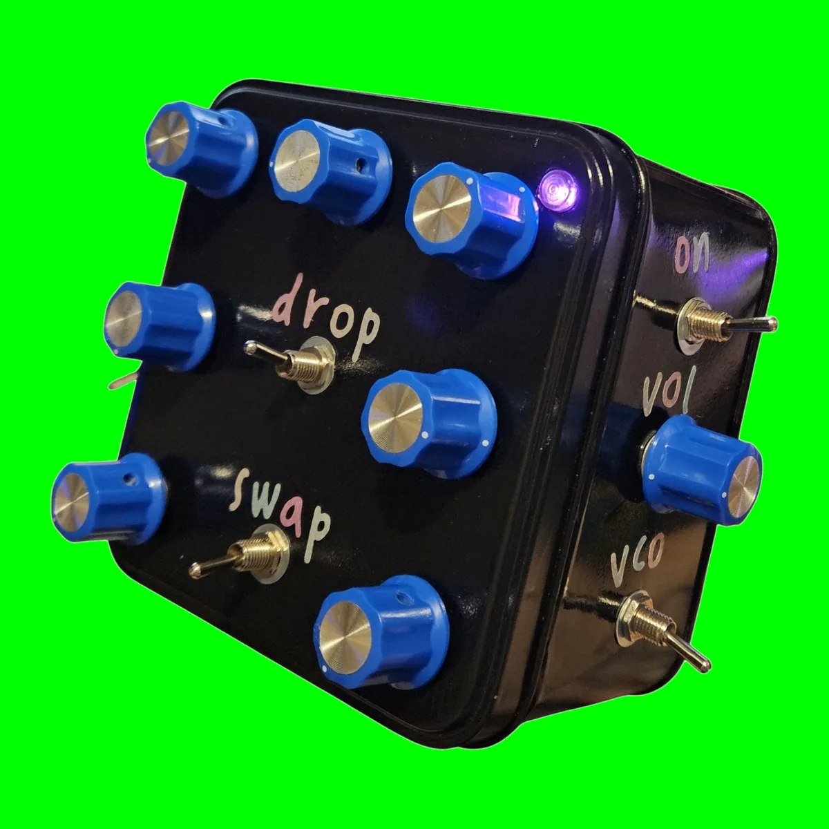 Bluebottle - a small square tin painted gloss black, seen from the front with one side showing: seven blue knobs on the lid, two toggles labeled drop and swap, a violet LED lit in the corner, and on the side a knob between two toggles labeled on, vol and vco