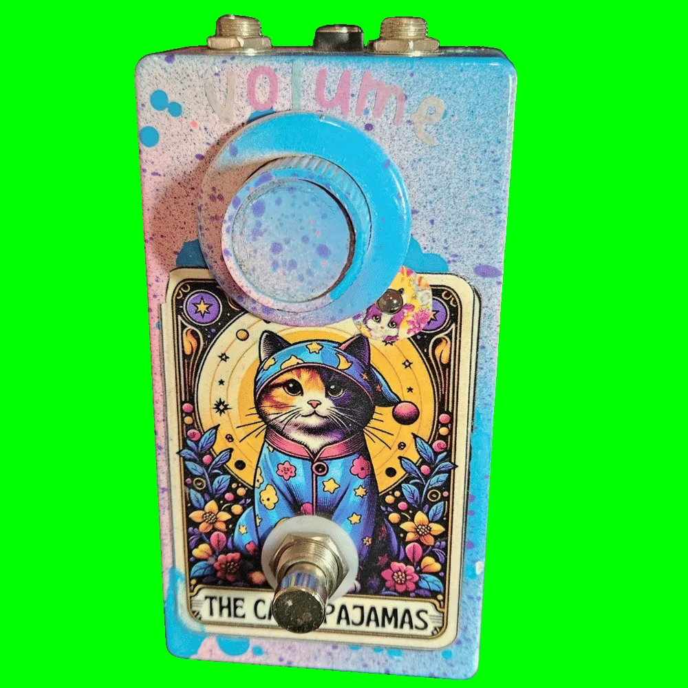 The Cat's Pajamas pedal - a tall enclosure in pastel pink, blue and lilac spatter with &ldquo;volume&rdquo; stencilled across the top in pink and white, one speckled pale-blue knob, and a tarot-card label of a calico cat in a star-patterned nightcap and pajamas seated against a golden crescent moon and ringed with flowers; a silver footswitch sits over the lower edge of the card, above the words THE CAT'S PAJAMAS